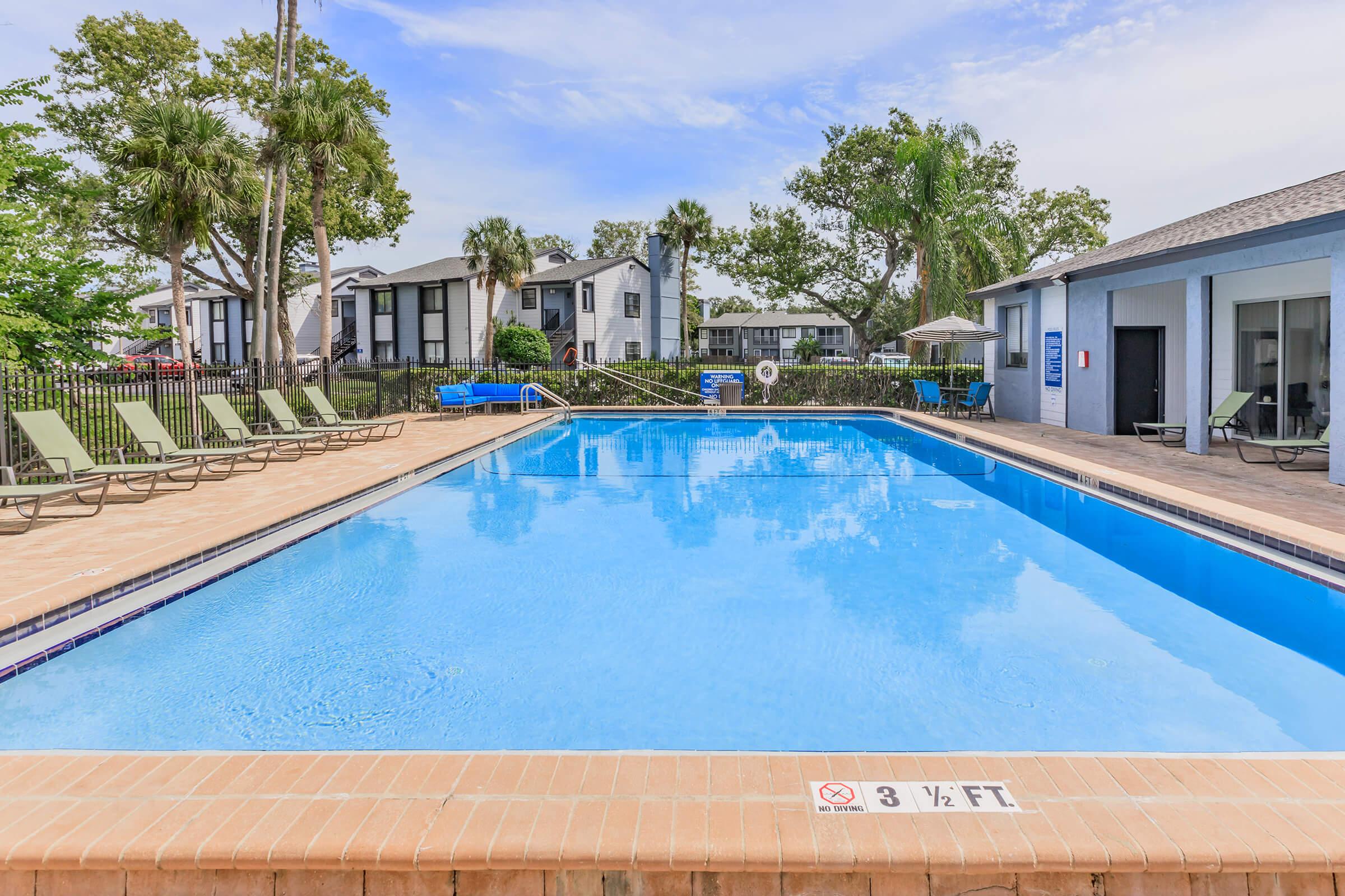 A clear blue swimming pool surrounded by lounge chairs and palm trees. The pool area features a paved deck and is located near residential buildings. Pool depth marker reads "3 ½ FT." The scene is sunny with a few clouds in the sky, creating a relaxing atmosphere.