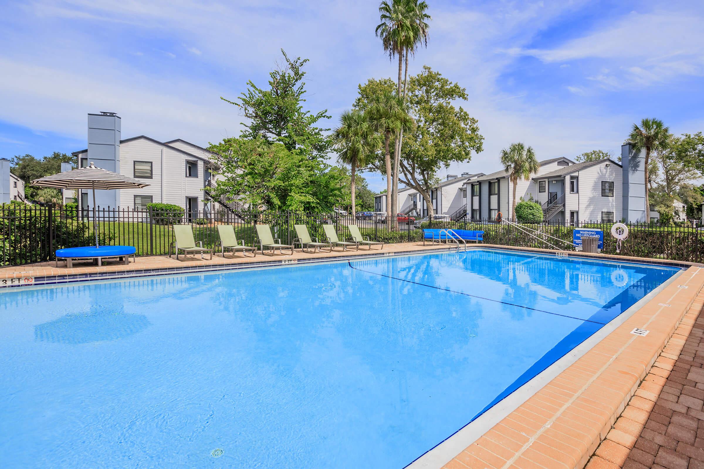 A clear blue swimming pool surrounded by green lounge chairs and lush trees. In the background, residential buildings are visible, adding to a serene atmosphere. A few umbrellas provide shade, creating an inviting outdoor space for relaxation and recreation.