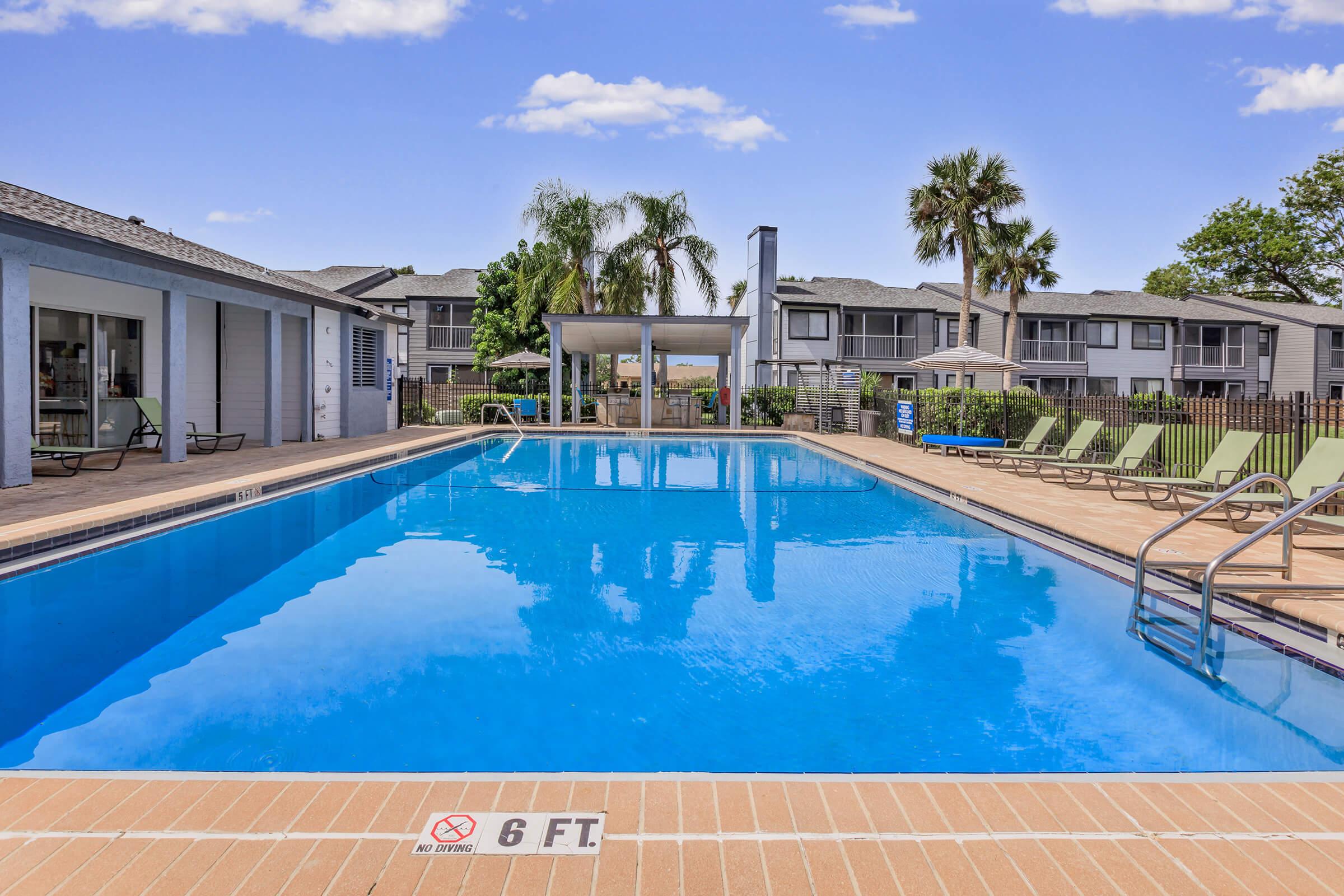 A sunny outdoor pool area featuring a clear blue pool surrounded by lounge chairs. In the background, there are palm trees and residential buildings. A safety sign indicates "6 FT" near the pool edge. The setting appears inviting and is perfect for relaxation.