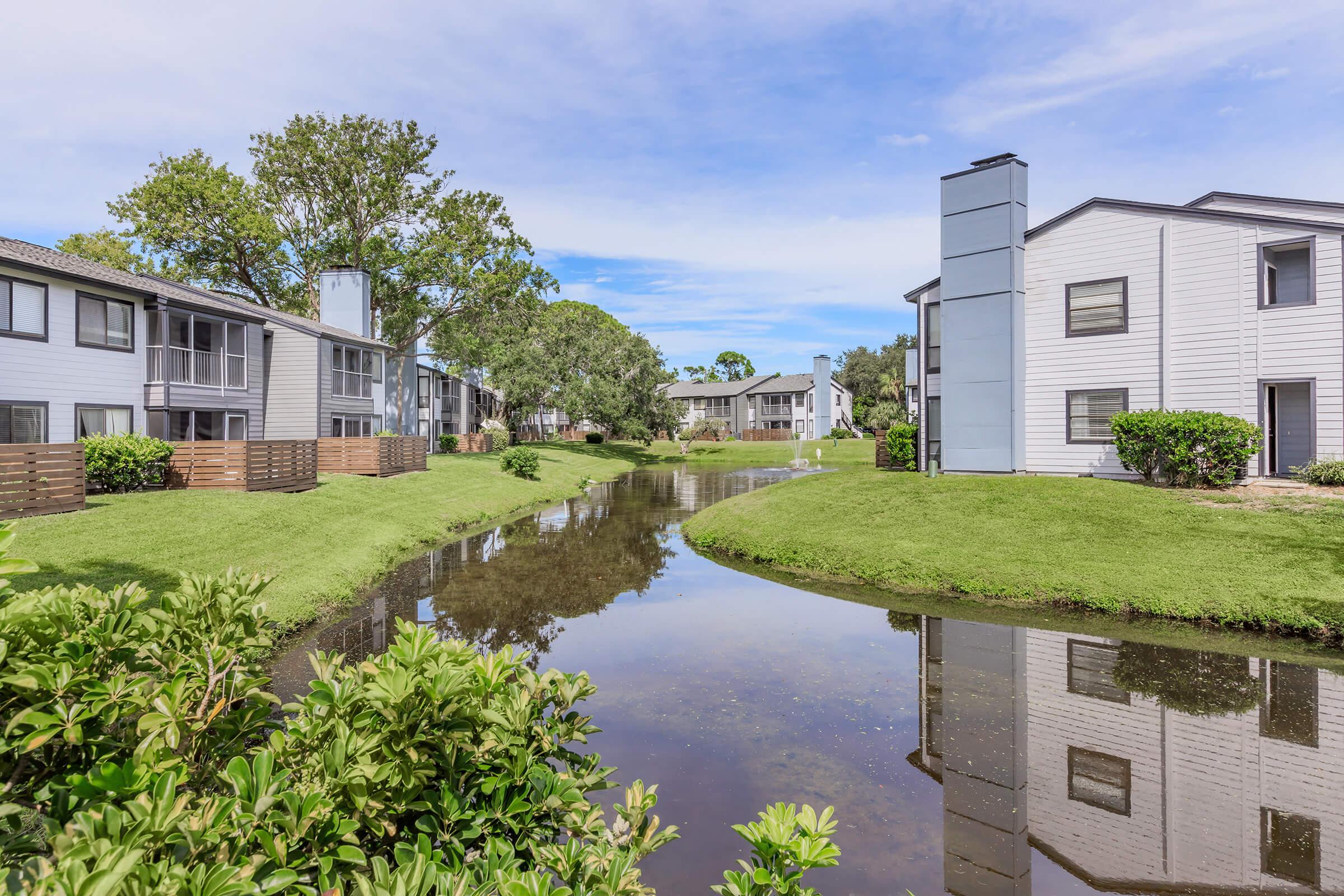 A scenic view of an apartment complex flanking a tranquil waterway. The reflection of the buildings and trees can be seen in the calm water, surrounded by well-maintained green lawns and shrubbery under a clear blue sky.