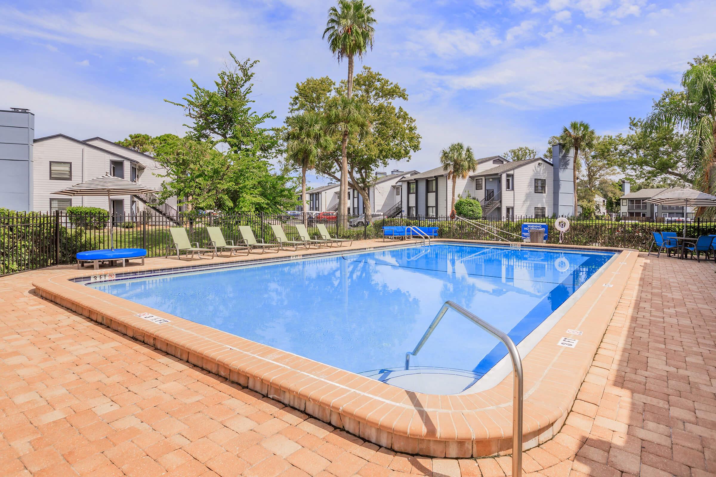 A clear blue swimming pool surrounded by brick paving and lounge chairs. Green trees and palm trees provide shade, while nearby buildings are visible in the background. Umbrellas are set up near the poolside, creating a relaxing outdoor atmosphere. The sky is bright with a few clouds.