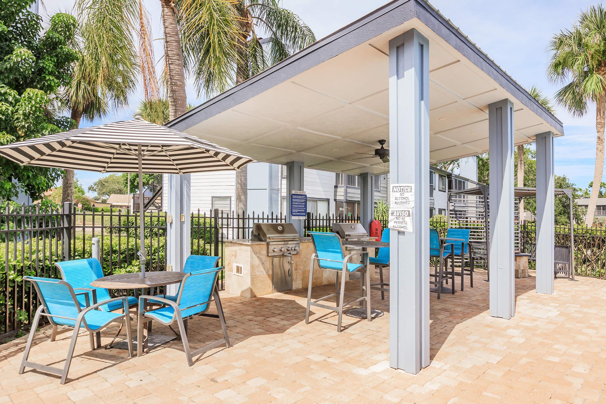 A shaded outdoor space featuring a grill station, a dining table with blue chairs, and a striped umbrella. The area is surrounded by greenery and has a fenced perimeter, creating a comfortable setting for gatherings or barbecues.