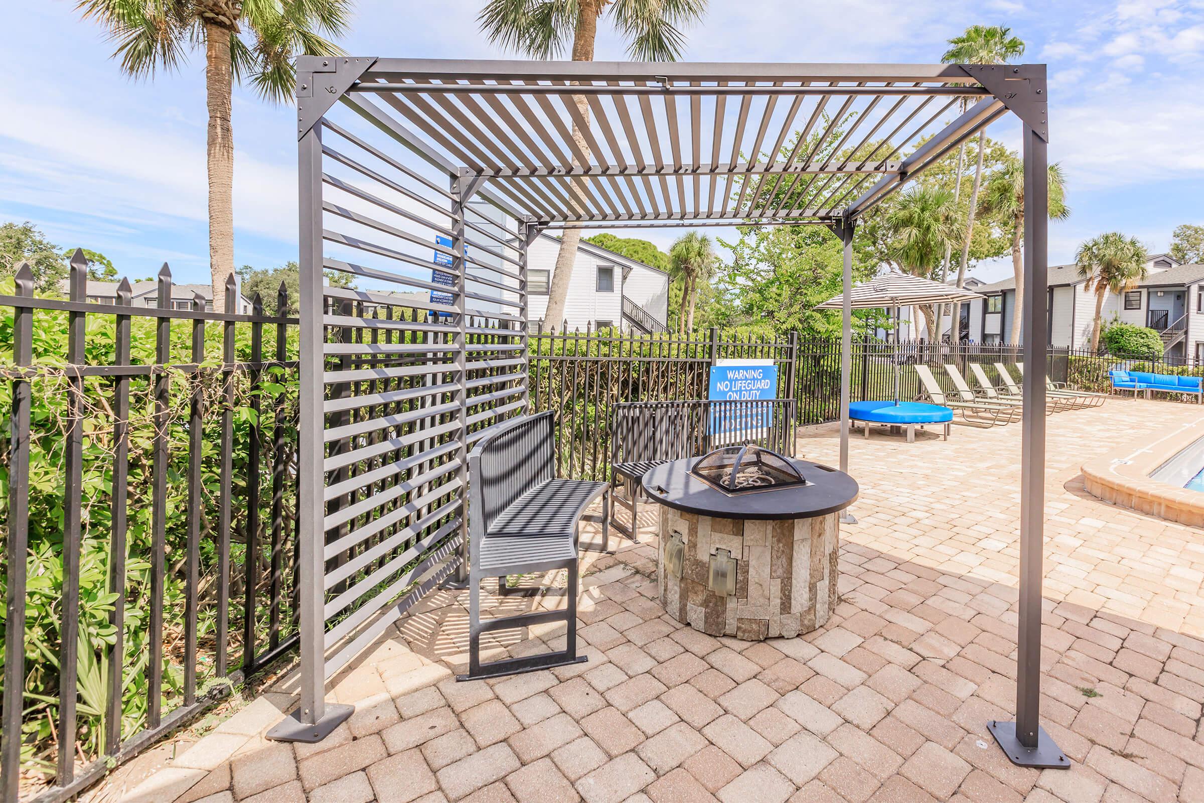 A shaded outdoor seating area with a metal frame and slatted roof, featuring a round fire pit at the center. Surrounding the area are palm trees and a fenced poolside setting, with lounge chairs in the background. Bright blue skies are visible above, enhancing the tranquil atmosphere.