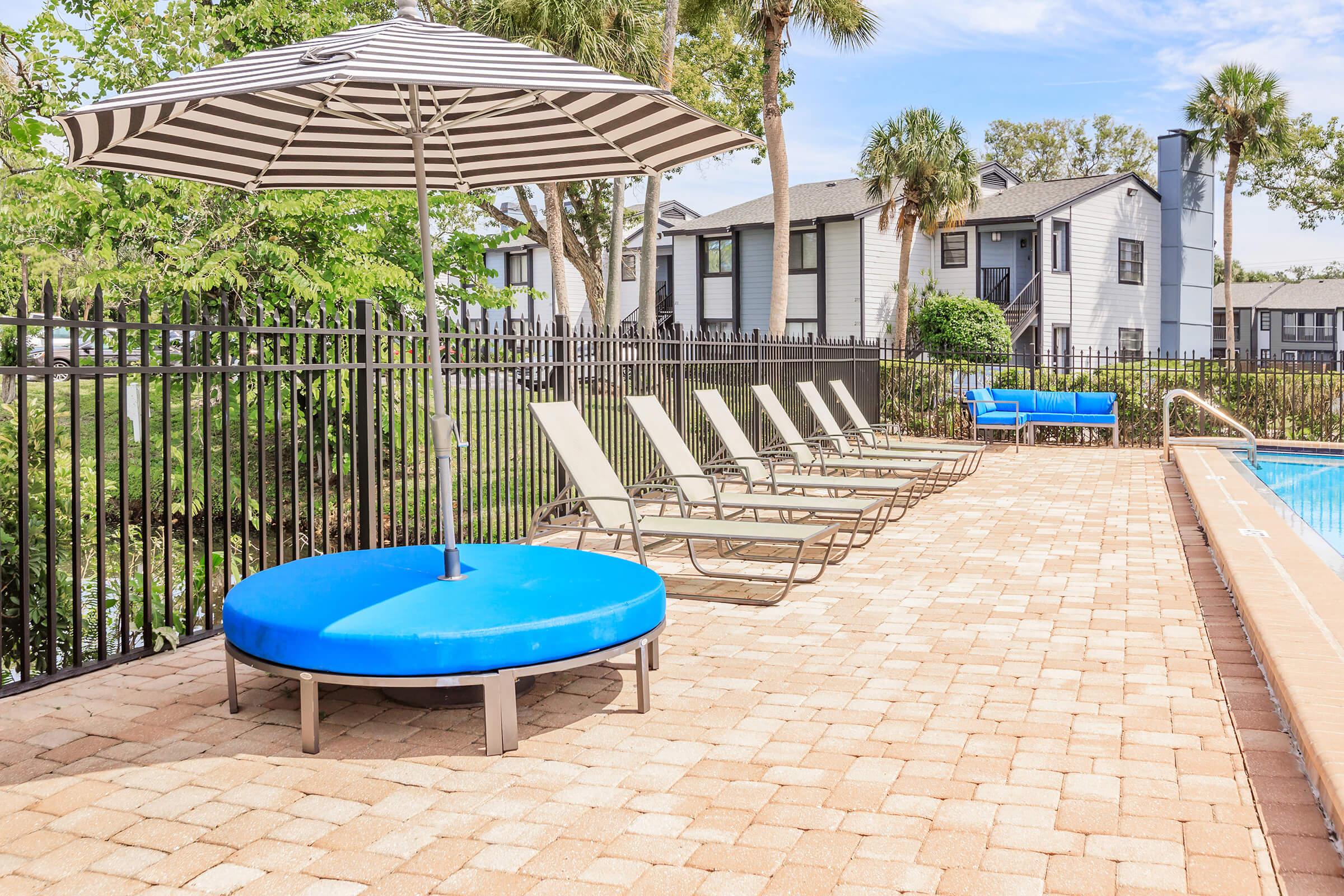 A sunny poolside scene featuring a circular blue lounge chair with a striped umbrella, several reclining chairs, and a swimming pool surrounded by a black fence and greenery. The background includes a few buildings with multiple windows.