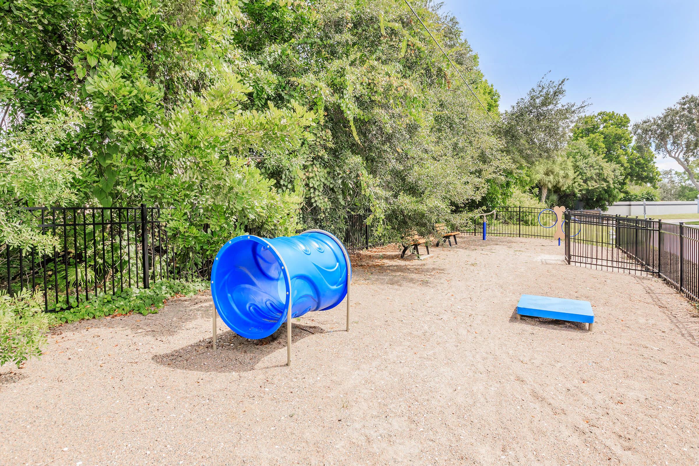 A blue playground barrel sits on sandy ground in a recreational area, surrounded by greenery and a black fence. There's a blue platform nearby, and in the background, a grassy field is visible. The scene is bright and sunny, ideal for outdoor activities.