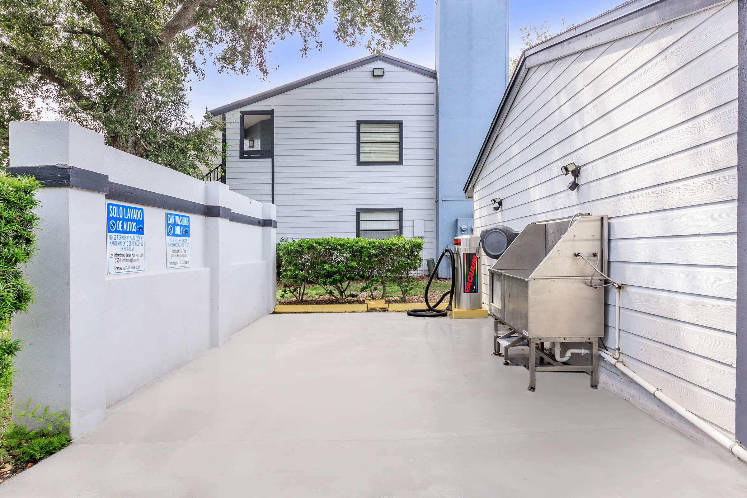 A clean, paved outdoor area beside a building featuring a metal utility cart, trash area, and signage on the walls. Green shrubs line the space, and there is a clear view of a two-story building with a light-colored exterior in the background.