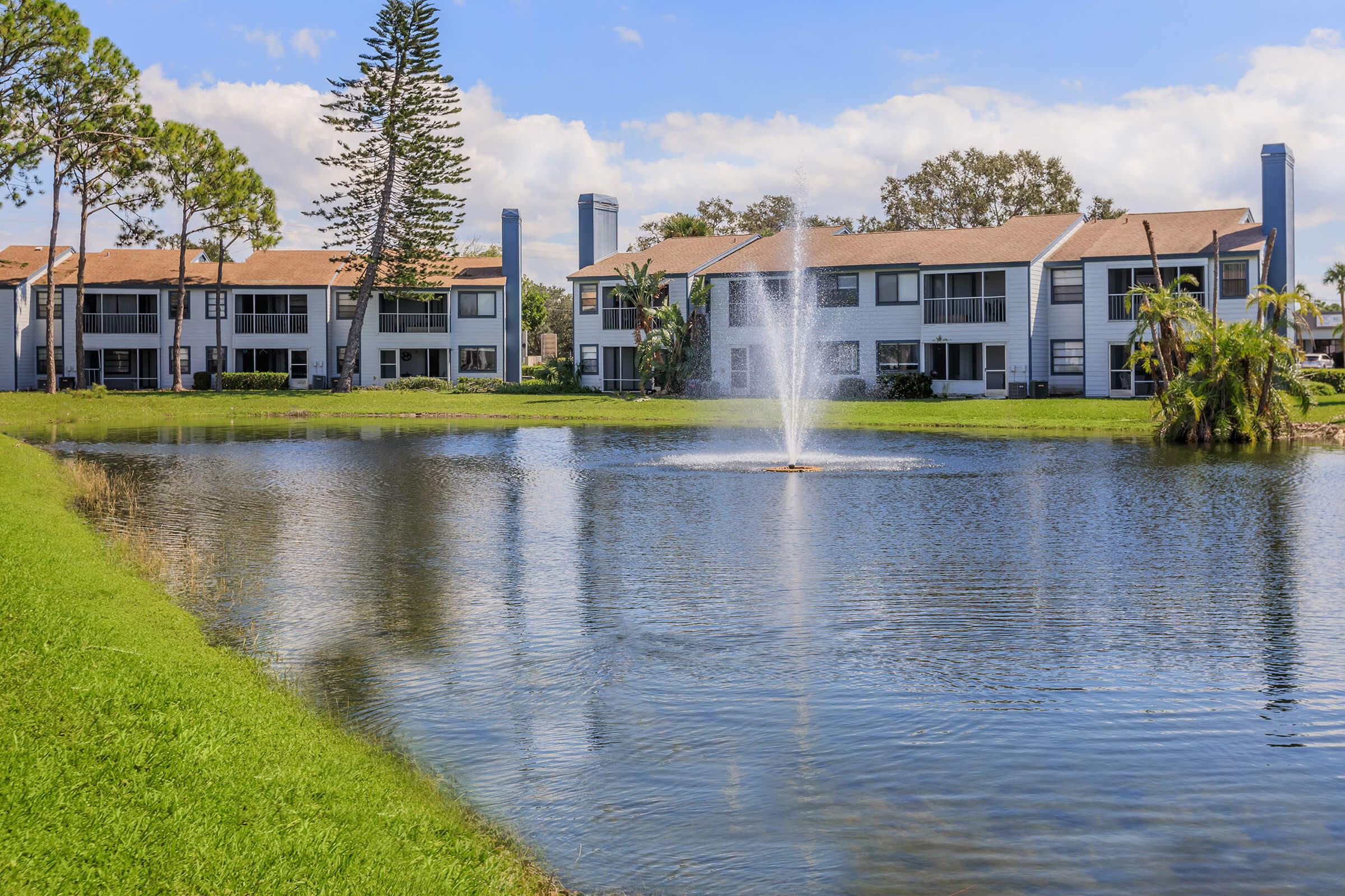 A peaceful lakeside scene featuring a fountain in the center of a calm pond. In the background, there are several modern apartment buildings surrounded by lush green grass and palm trees, with a clear blue sky and fluffy white clouds above.
