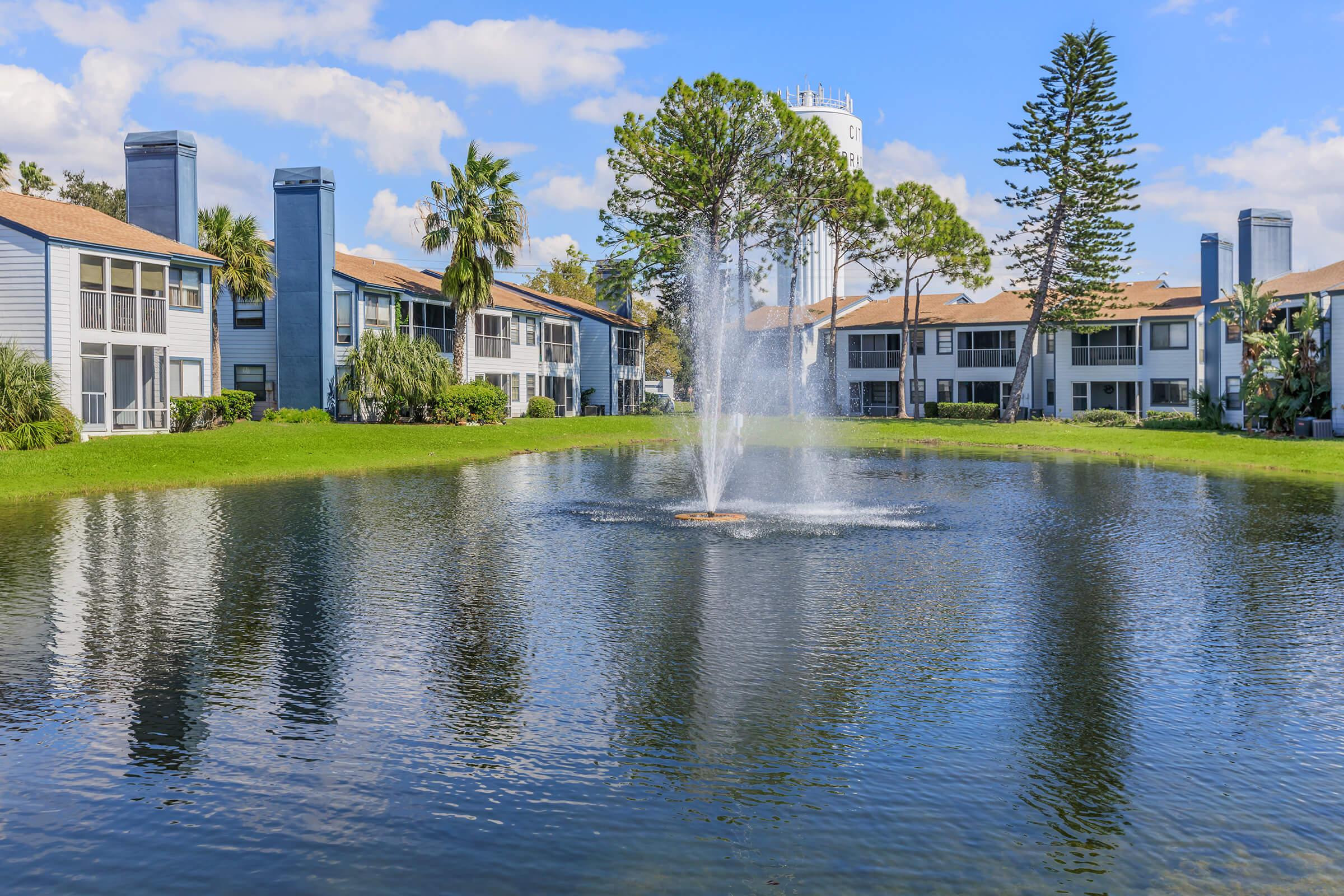 A serene landscape featuring a calm pond with a fountain, surrounded by green grass and palm trees. In the background, there are two-story apartment buildings with balconies. The sky is partly cloudy, creating a peaceful atmosphere.