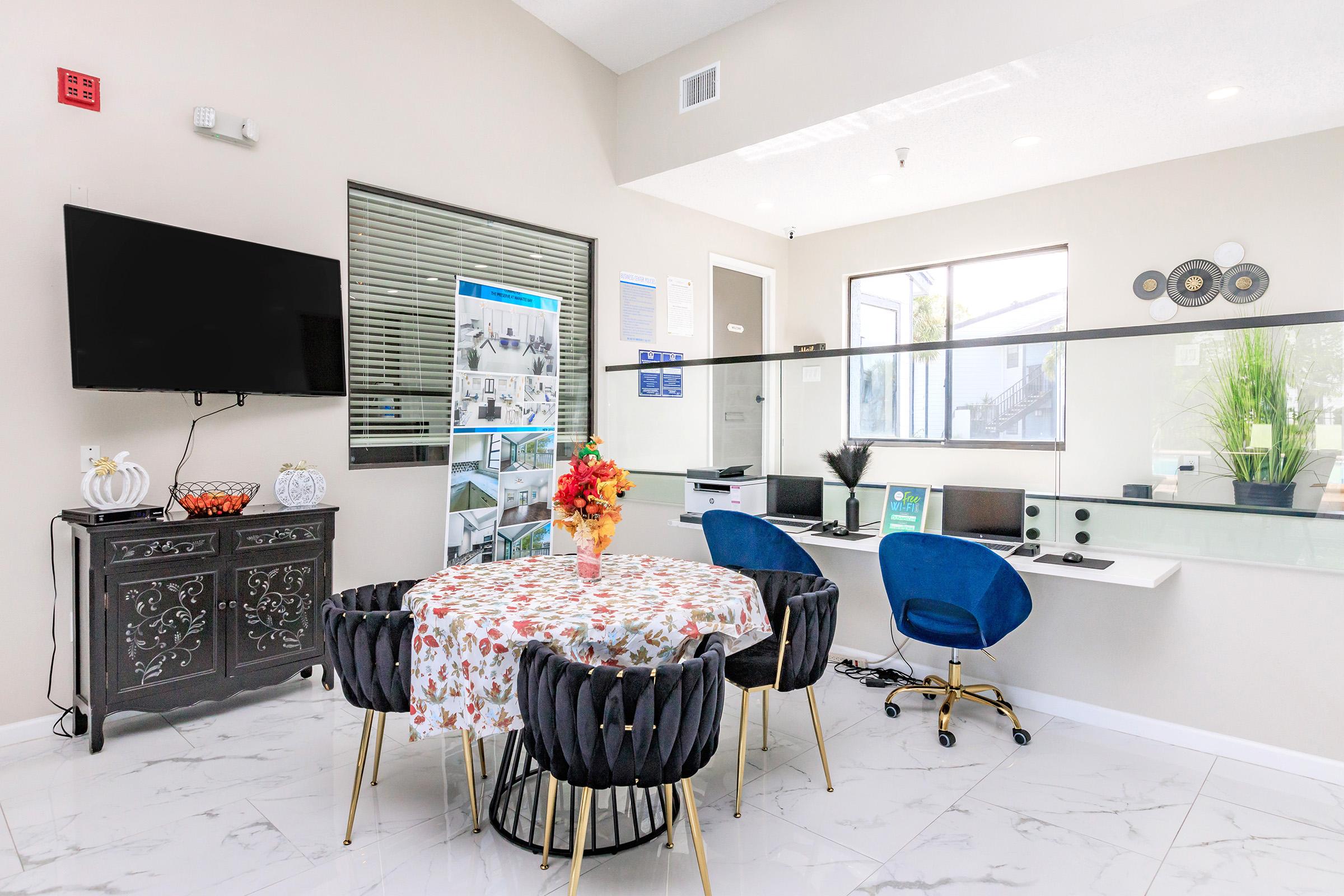 A bright and modern reception area featuring a round table with a floral centerpiece surrounded by stylish chairs. In the background, there's a glass partition with workstations that include computers and speakers, along with a wall-mounted flat-screen TV and decorative wall art.