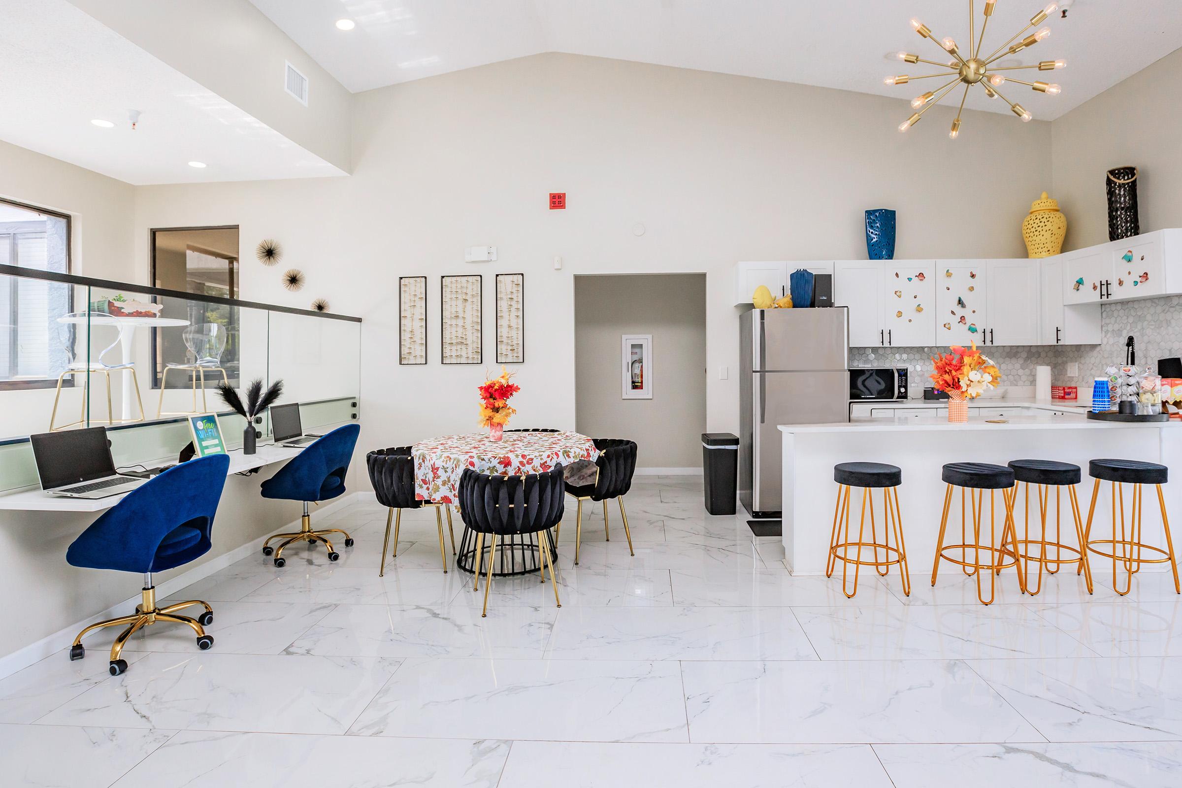 A modern open-concept kitchen and dining area featuring a round table with floral tablecloth, high-backed blue chairs, and a sleek kitchen island with bar stools. The room has bright lighting, white marble flooring, and elegant decor including wall art and a stylish chandelier. Workstations with computers are visible in the background.