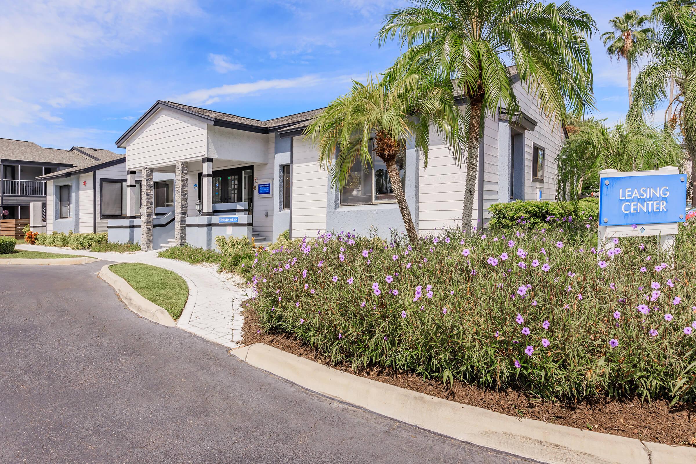 A leasing center building surrounded by lush greenery and blooming flowers. The entrance is visible with a blue sign indicating "Leasing Center." The pathway leading up to the building is lined with palm trees and well-maintained landscaping, creating an inviting atmosphere.