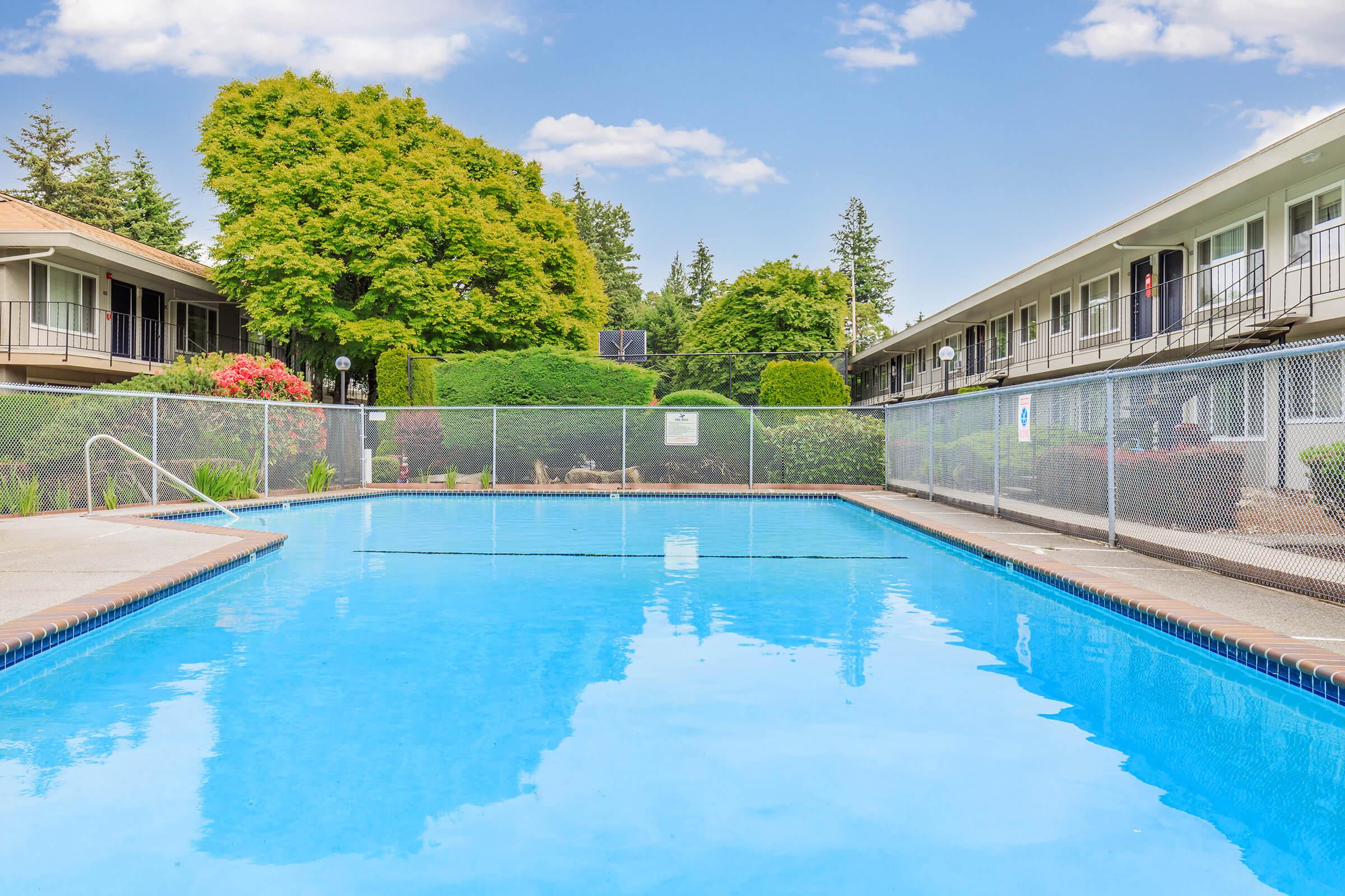 A clear blue swimming pool surrounded by a protective fence, with lush green hedges and trees in the background. The pool area is set against a backdrop of a low-rise building, and the sky is bright with scattered clouds.