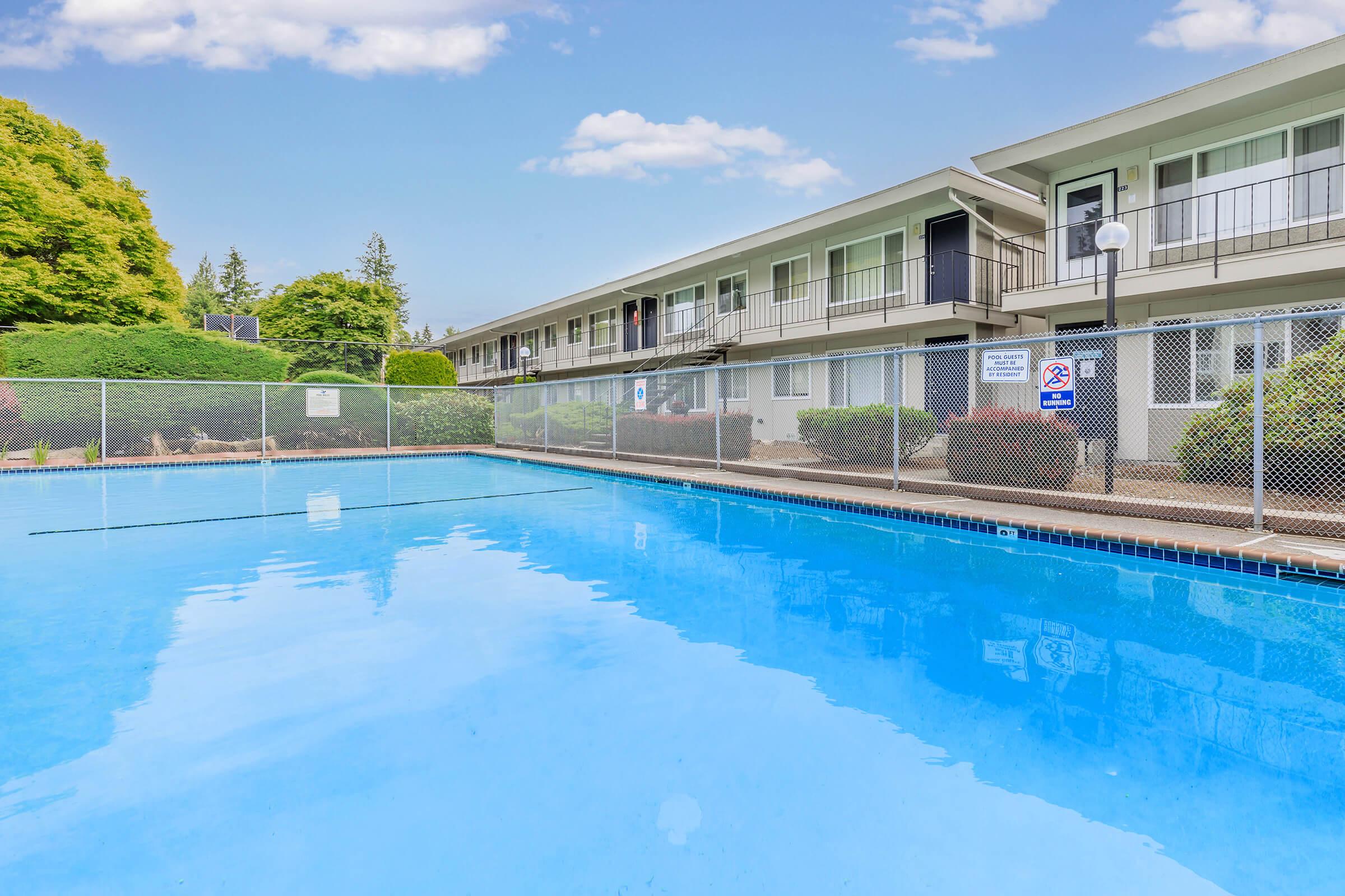A clear swimming pool with a chain-link fence around it, surrounded by lush greenery. In the background, there are two-story buildings with balconies. The sky is blue with a few clouds. Signs are visible near the pool area, indicating regulations.