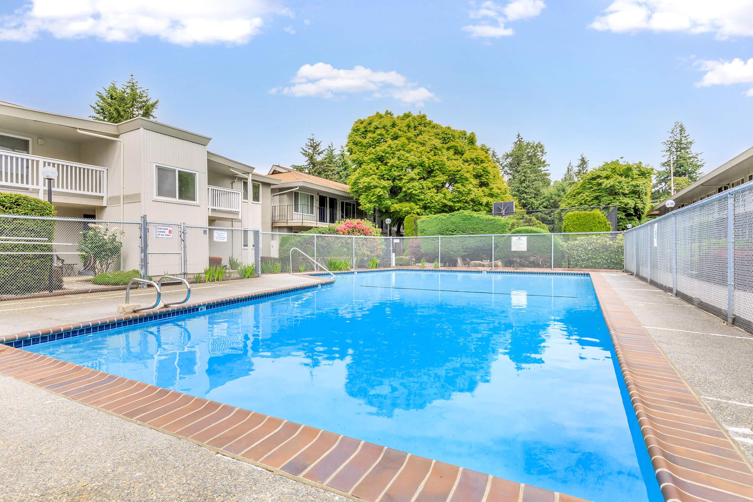 A bright outdoor swimming pool surrounded by landscaped greenery and residential buildings. The pool has clear blue water, a diving board, and is enclosed by a fence. Nearby, there are trees and shrubs adding to the peaceful atmosphere under a clear blue sky.