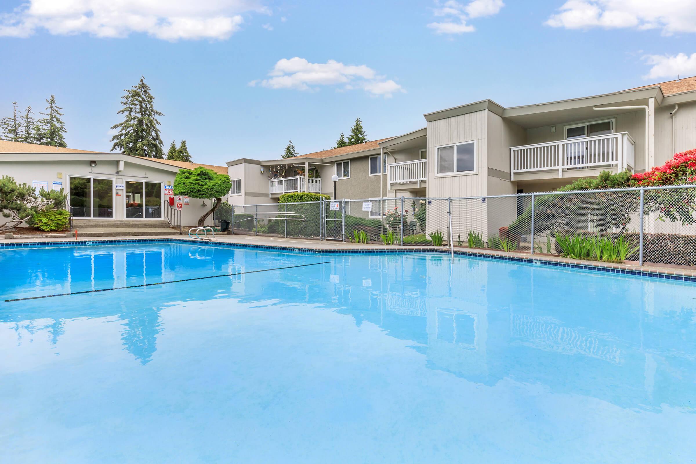 A clean, inviting swimming pool surrounded by lush greenery and well-kept landscaping, with residential buildings in the background. The sky is clear with a few clouds, and there are lounge chairs by the poolside.