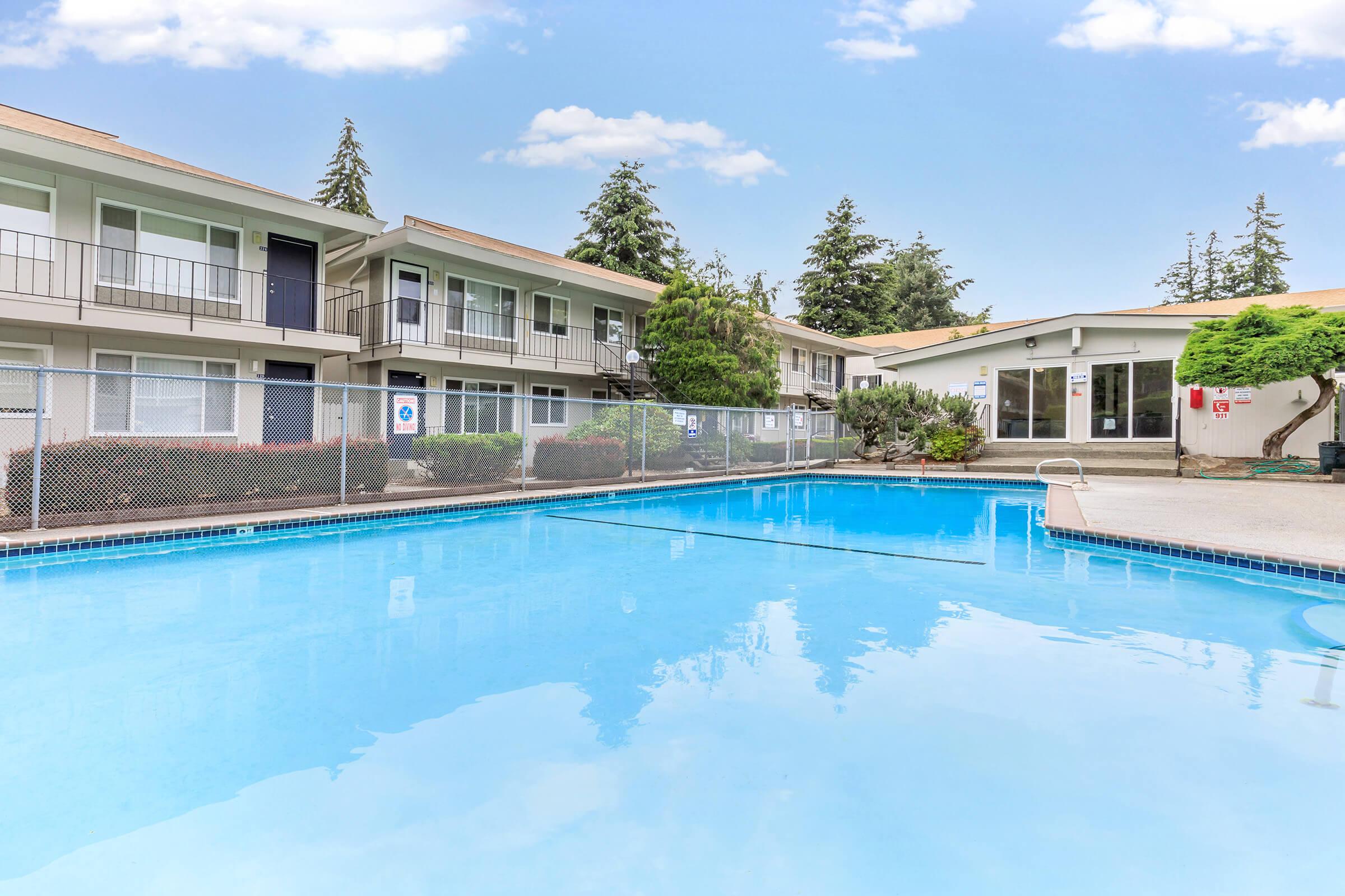 A clear blue swimming pool surrounded by a fenced area, with two multi-story buildings in the background. Lush green trees and shrubs are visible near the pool area. The sky is bright with a few clouds, enhancing the overall inviting atmosphere of the setting.