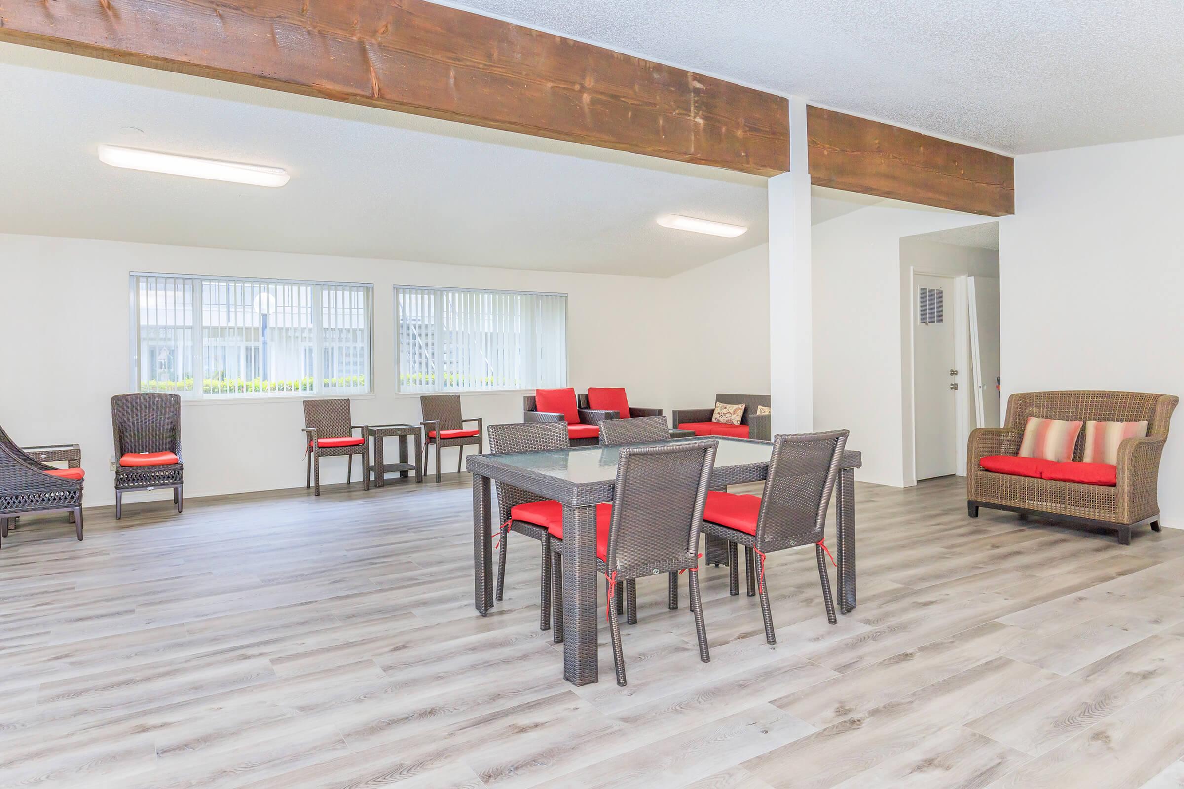 A spacious interior room featuring a modern design with wooden beams and large windows. There are several seating areas, including a sofa and dining table set with red-accented chairs and a dark wicker style. The floor is made of light wood, creating a bright and inviting atmosphere.