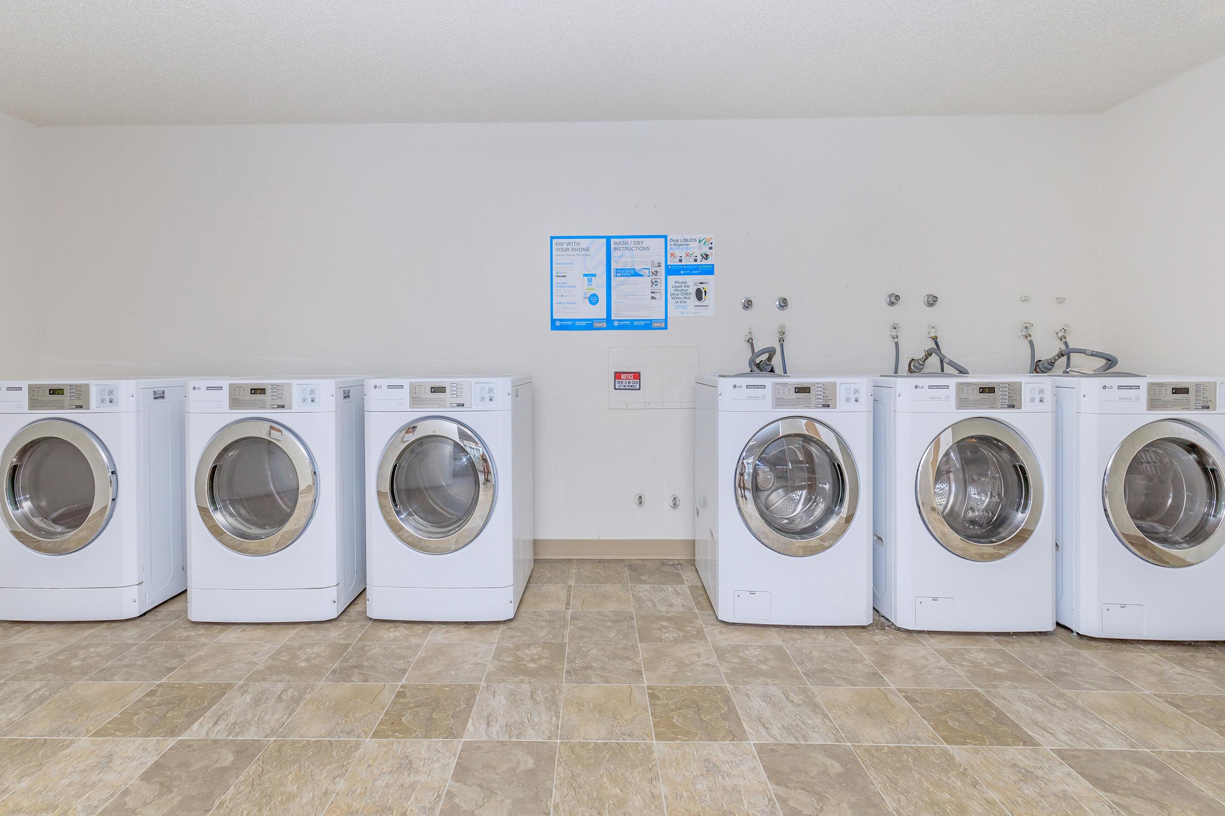 A row of six white washing machines lined up against a wall in a laundry room. They are front-loading models, with clear glass doors. The floor features tiled patterns in shades of brown and beige. A bulletin board with laundry instructions and information is mounted on the wall above the machines.