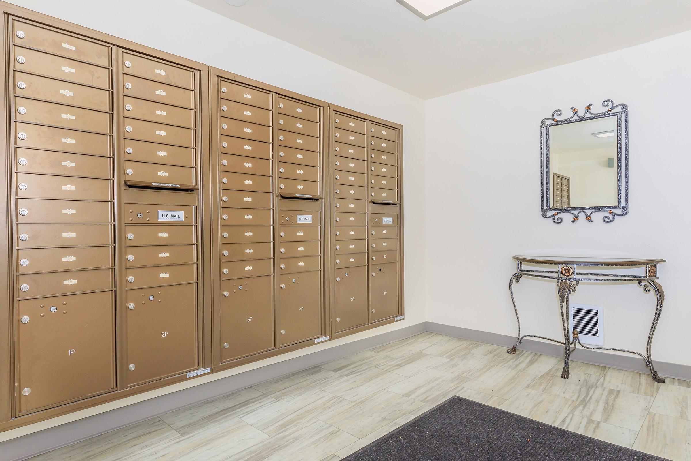 A well-lit hallway featuring several bronze mailboxes mounted on the wall. There's a decorative mirror and a small, ornate table against the wall, with a dark mat on the floor. The space has a clean and modern appearance, with neutral wall colors and tiled flooring.
