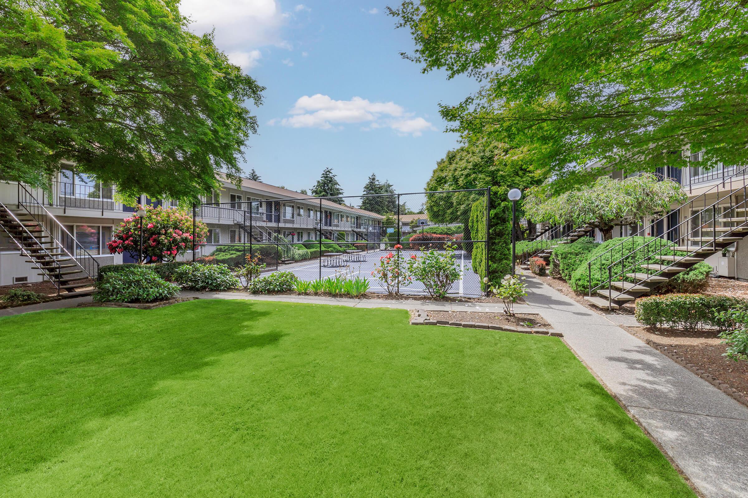 A well-maintained courtyard featuring lush green grass and well-kept plants, surrounded by a two-story building. There are stairs leading to balconies on either side and a swimming pool in the center, with clear blue skies and a few clouds above, creating a serene and inviting atmosphere.