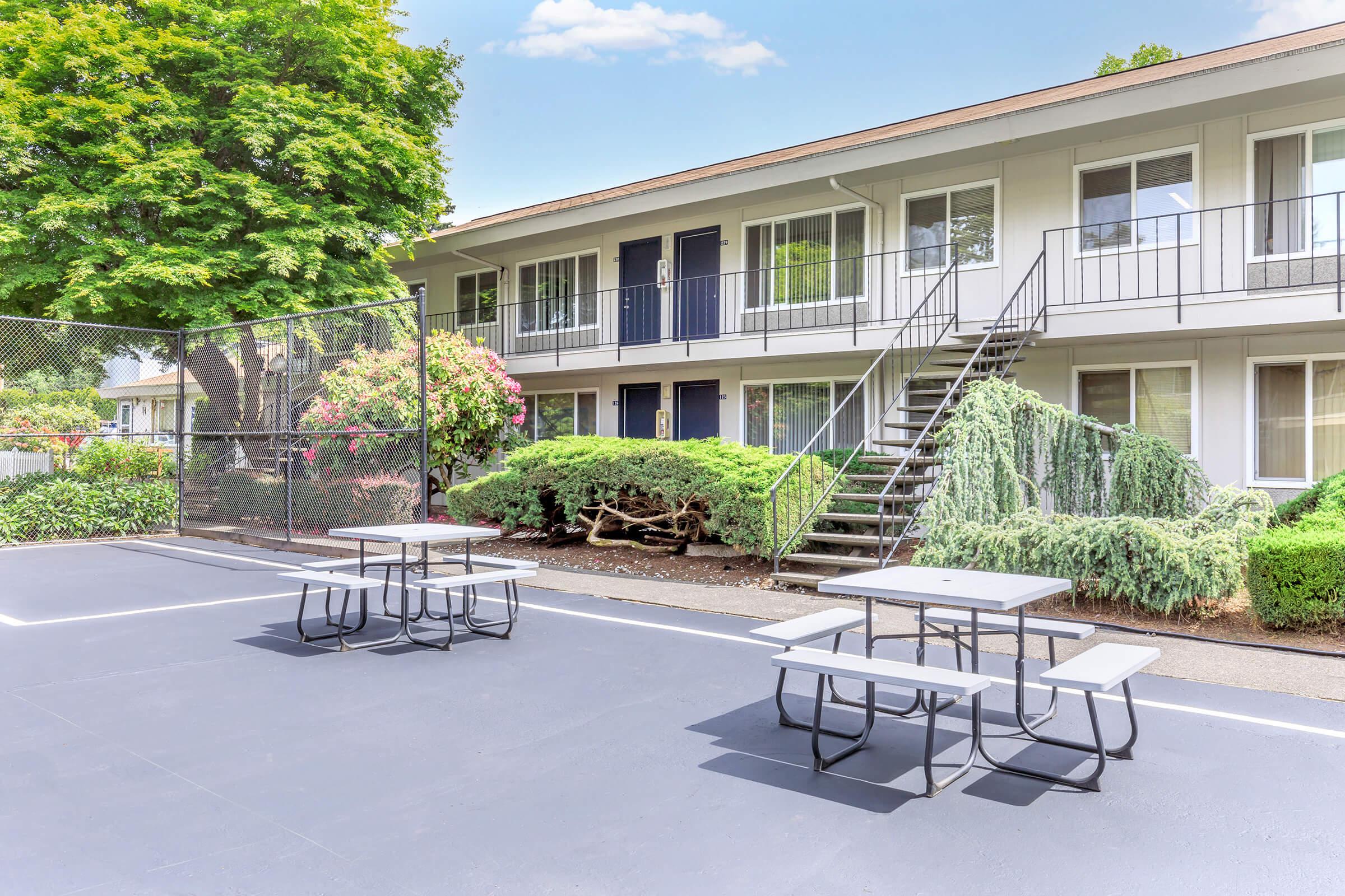 A view of a multi-unit apartment building with a fenced basketball court in the foreground. Several picnic tables are arranged on the court surface. Lush greenery and flowering plants surround the building, and a staircase leads to the upper level of the apartments. The sky is clear and blue.