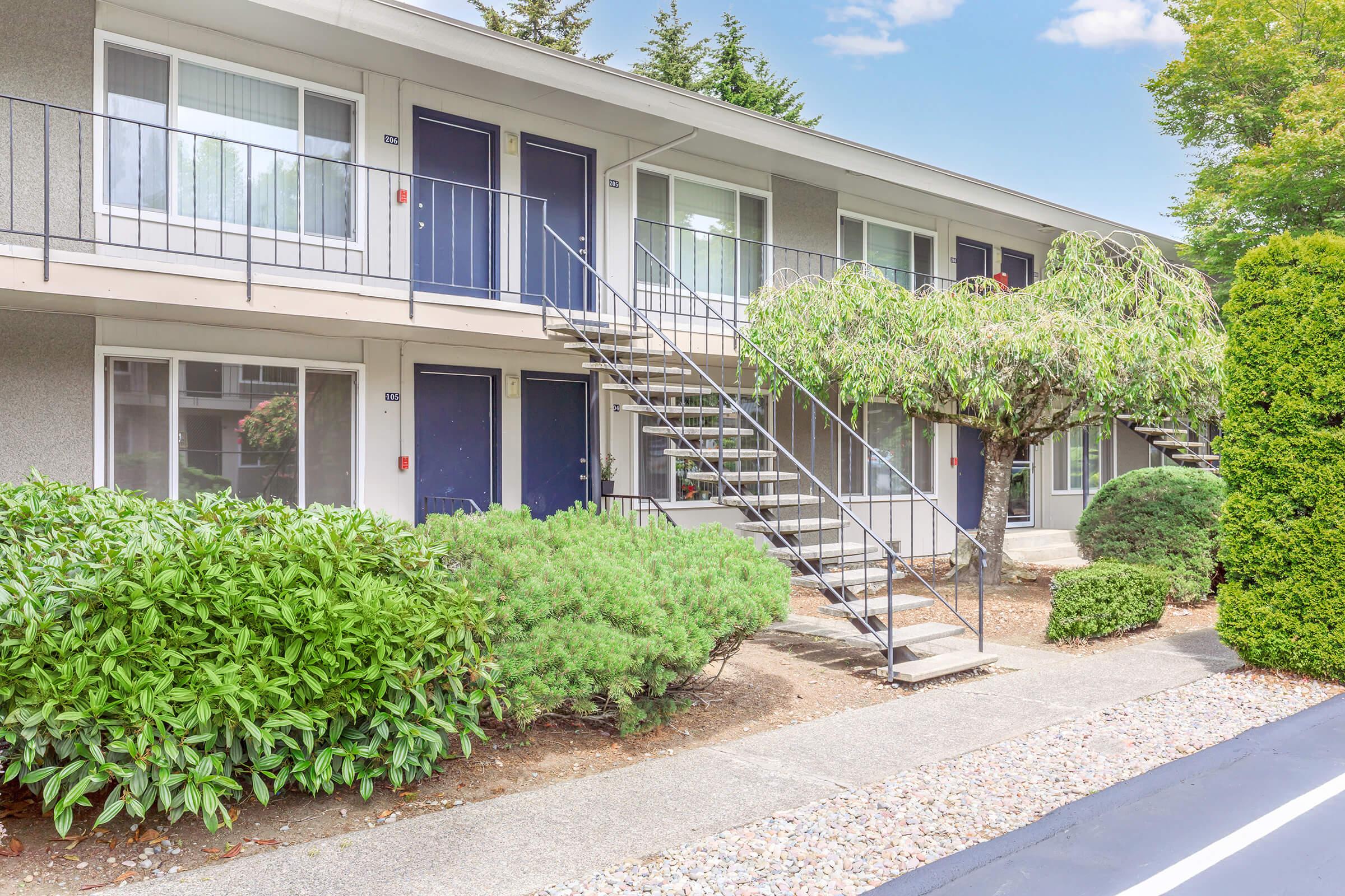 A view of a two-story apartment building featuring a staircase leading to the second floor. The building has multiple entrances, surrounded by well-maintained landscaping including shrubs and a small tree. The setting is bright and inviting, with clear skies in the background.