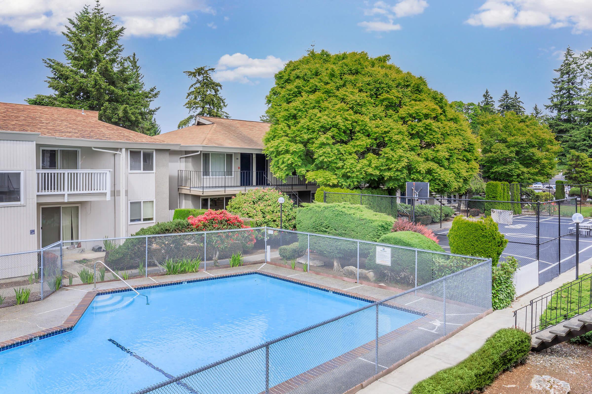 A view of an outdoor swimming pool surrounded by greenery and well-maintained landscaping, with residential buildings in the background. The area is sunny, featuring trees, colorful flowers, and a basketball court in the distance. A chain-link fence encloses the pool area for safety.