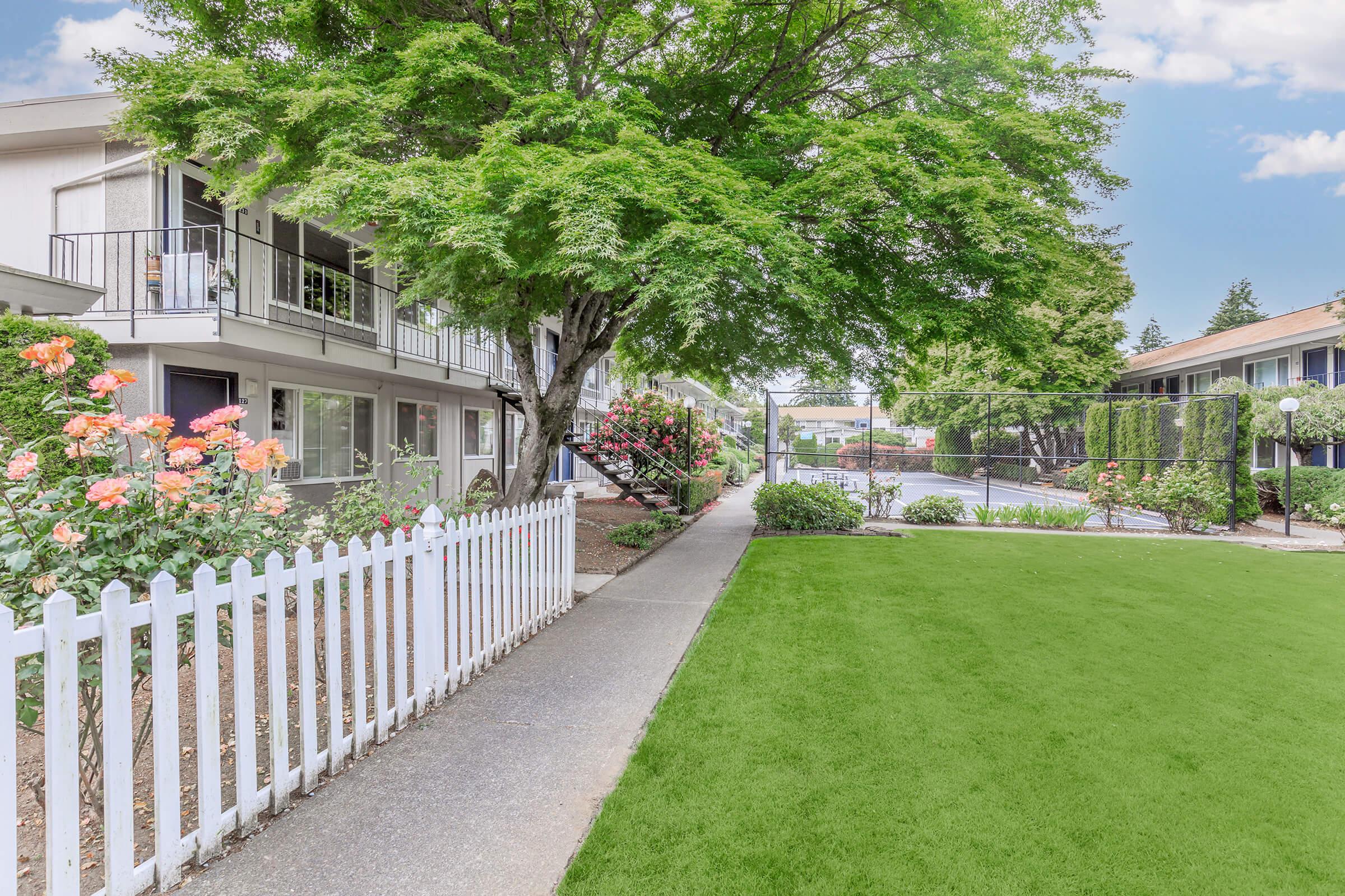 A well-maintained pathway leads through a landscaped area featuring vibrant flowers and lush green grass. On one side, there are two-story apartment buildings with balconies, while a swimming pool area is visible in the background, framed by hedges and trees under a clear blue sky.