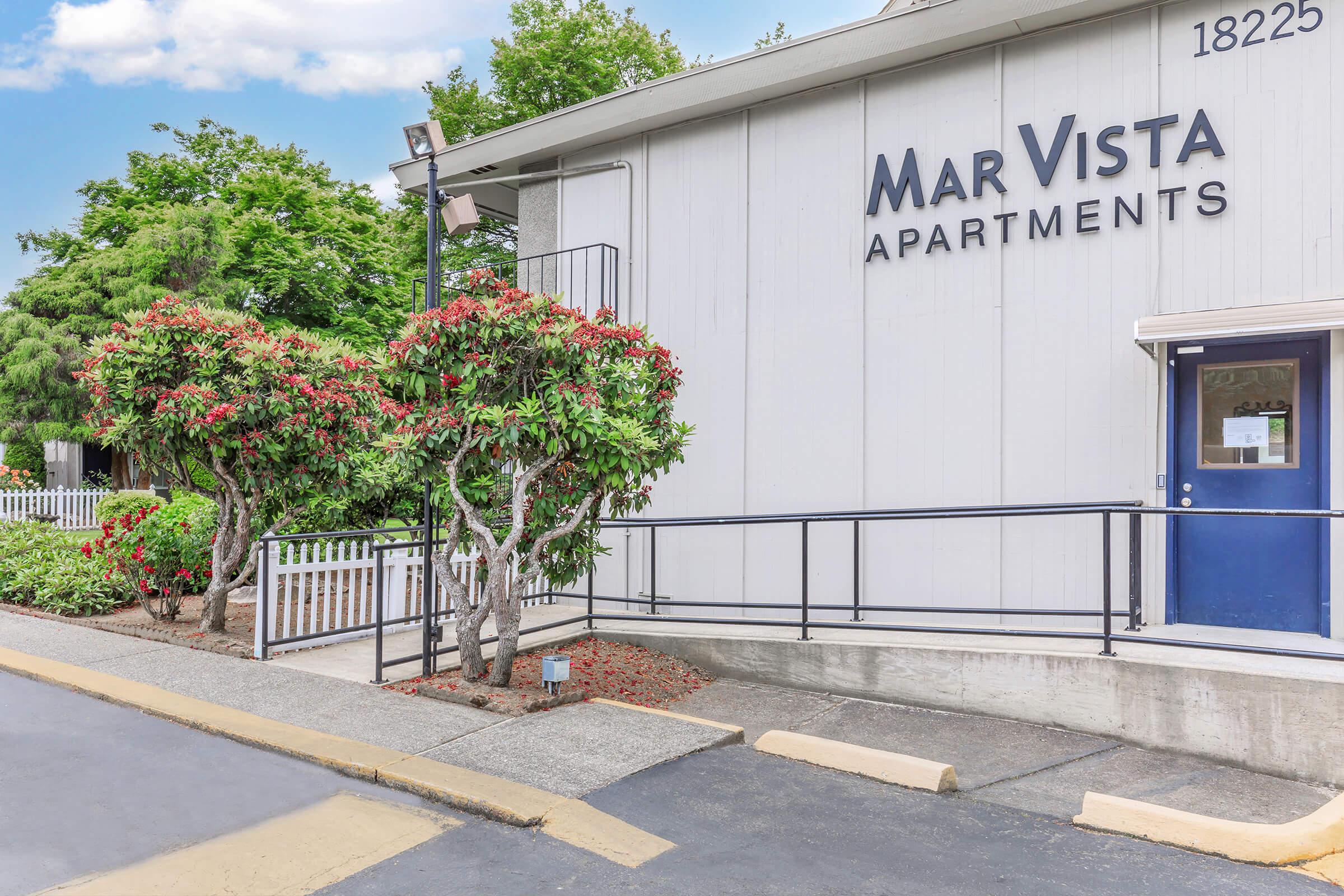 Exterior view of Mar Vista Apartments featuring a white building with the name prominently displayed. There are well-maintained shrubbery and trees in front, along with a ramp leading to the entrance. A nearby fence and a blue door complete the scene, set against a clear sky.