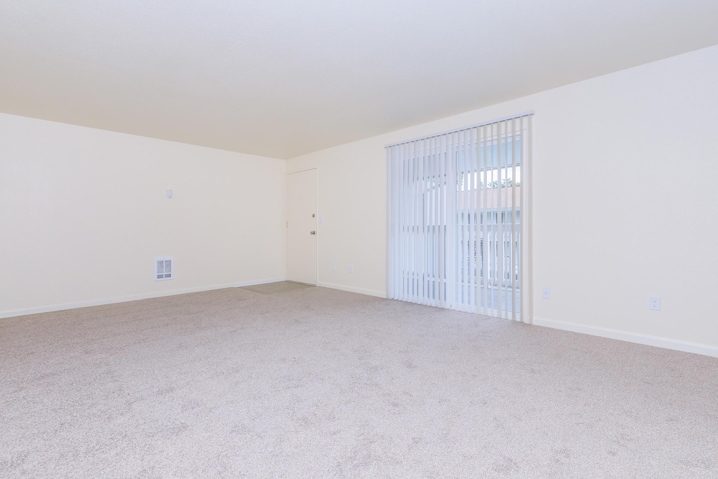 A spacious interior of an empty room with beige walls and light-colored carpet. The room features a sliding glass door with vertical blinds, leading to a balcony. Natural light fills the space, showcasing its openness and neutral decor.
