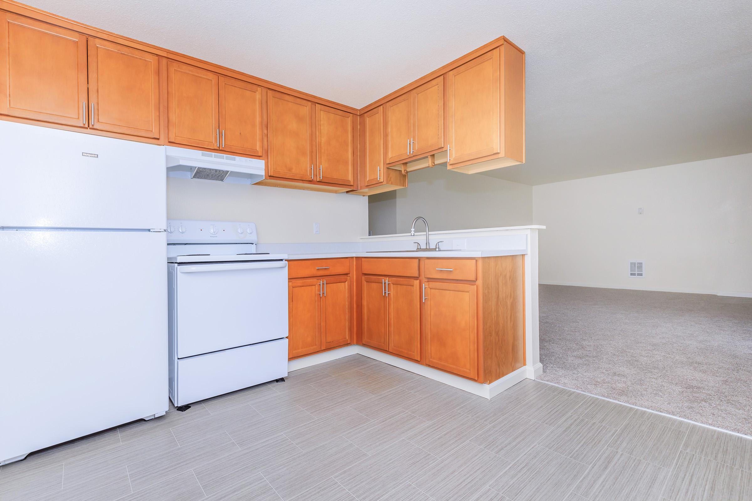 A modern kitchen with wooden cabinets, a white refrigerator, and a white stove. The countertop is clean and features a sink. The area has neutral-colored walls and a carpeted living space visible in the background.