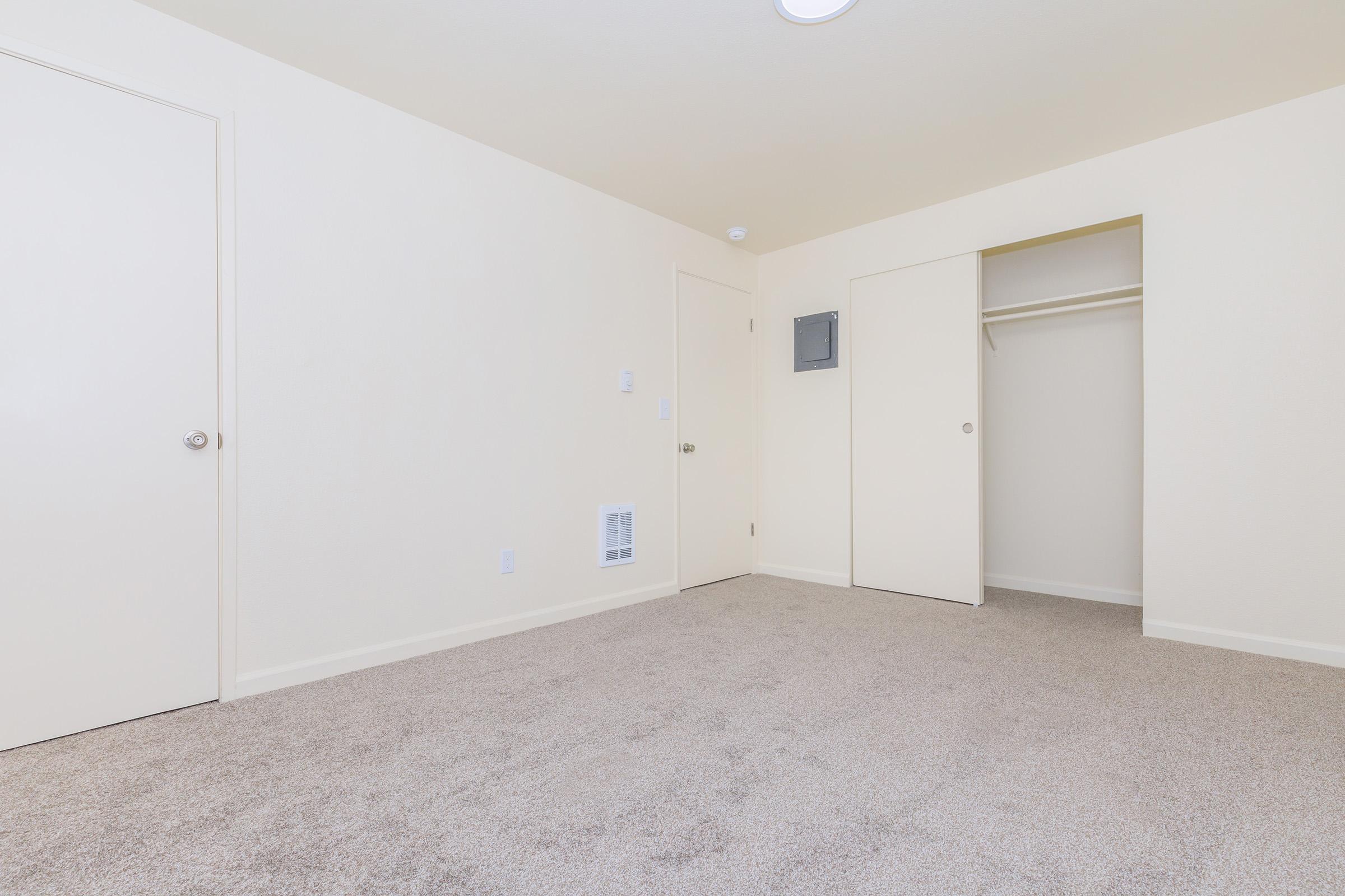 Empty room with light-colored walls and plush carpet. One door leads to another area, and there is a closet with sliding doors. A small access panel is visible on the wall. The space has a bright, neutral atmosphere, suitable for various uses.