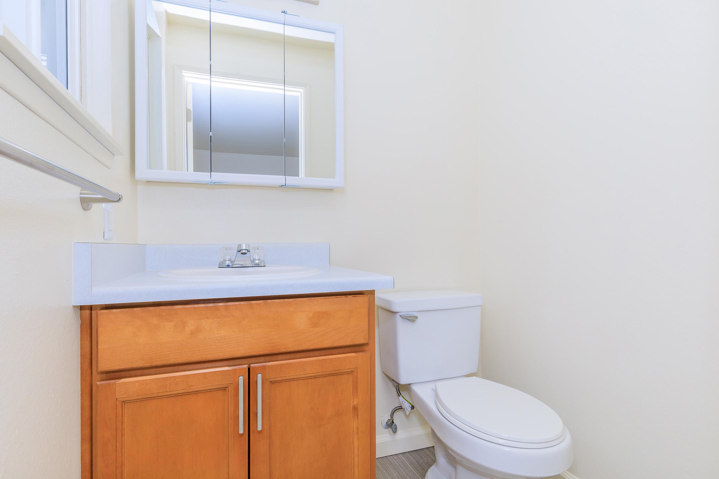 A clean, modern bathroom featuring a white toilet, a wooden vanity with a sink, and a wall-mounted mirror. The walls are painted in a light color, creating a bright and airy atmosphere. The flooring is neutral-toned, adding to the overall simplicity and elegance of the space.