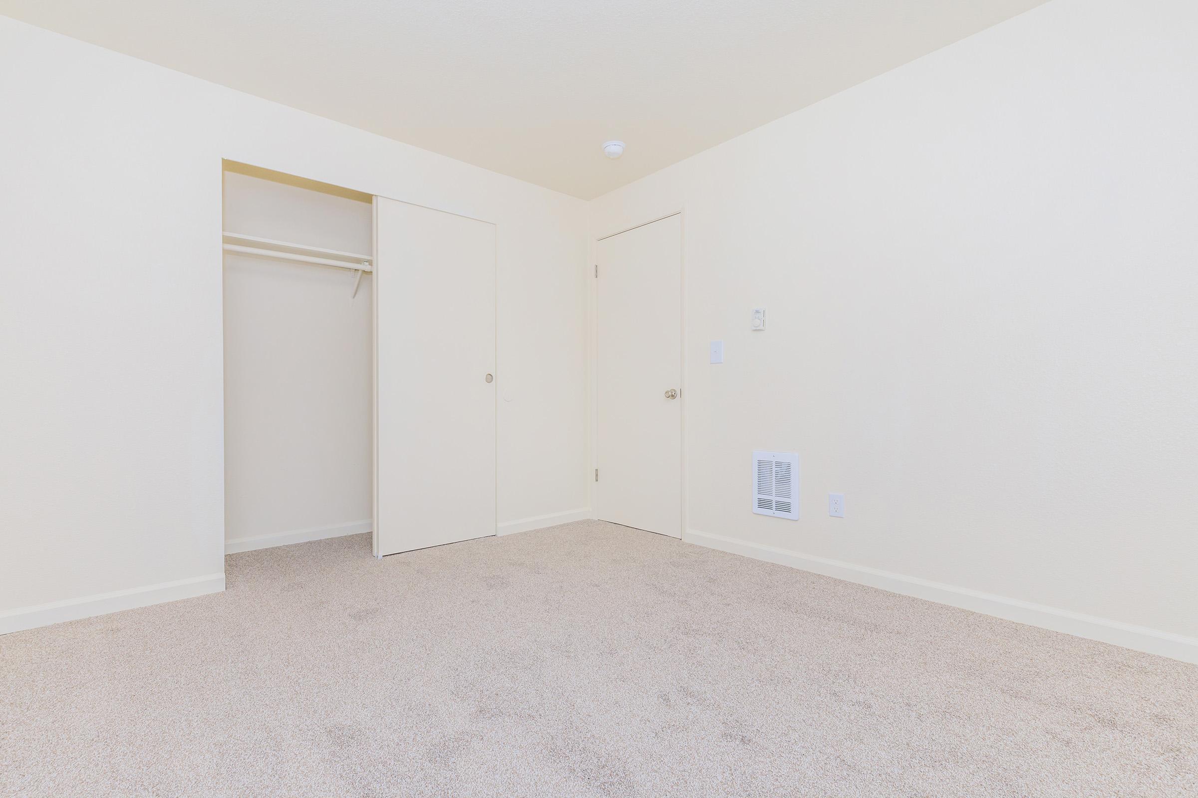 A simple, empty room with beige walls and carpet. It features a small closet with a sliding door on the left and a standard door on the right. The room is well-lit and unadorned, creating a minimalist appearance suitable for various uses.