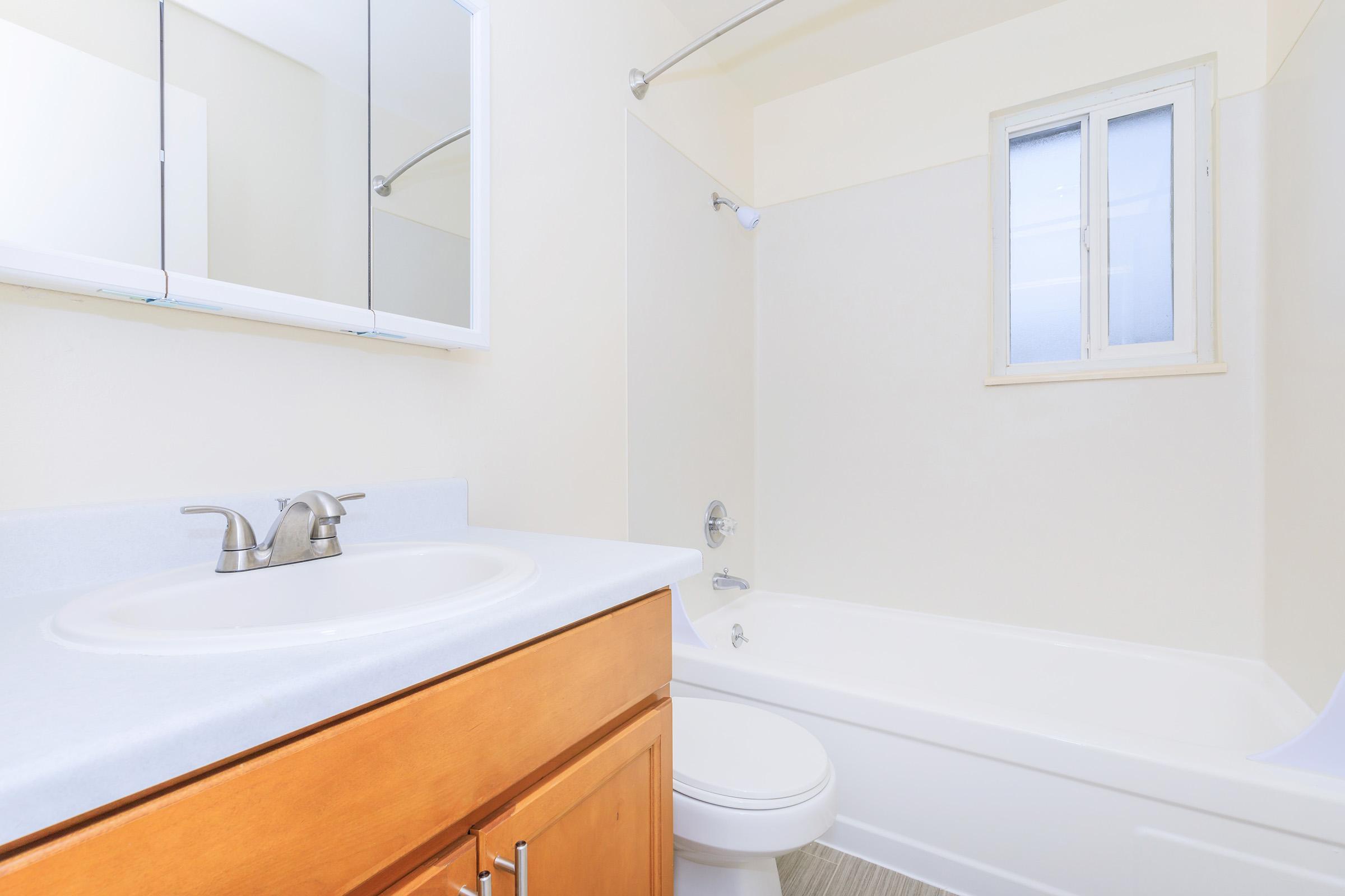 A bright, modern bathroom featuring a white countertop sink with a rectangular mirror above, a bathtub with a shower head, and a small window providing natural light. The walls are painted a soft light color, and there’s a simple wooden cabinet beneath the sink.