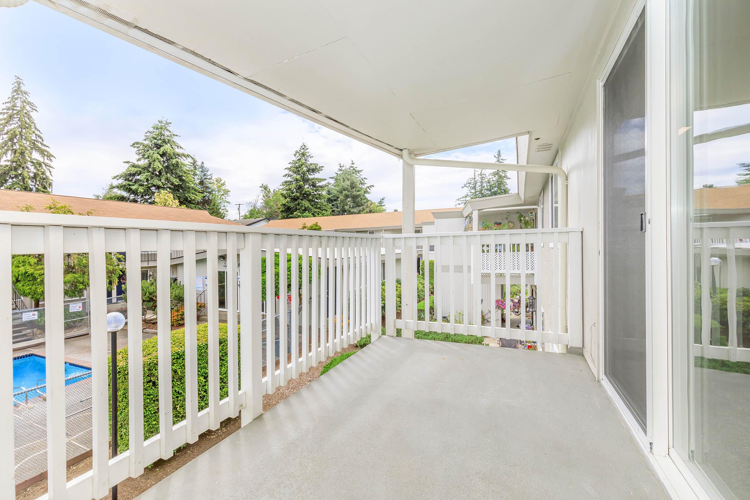 A spacious balcony with a white railing, overlooking a green courtyard and a swimming pool. The sky is partly cloudy, and tall trees are visible in the background, providing a serene atmosphere. The balcony floor is plain and offers a view of the surrounding area.