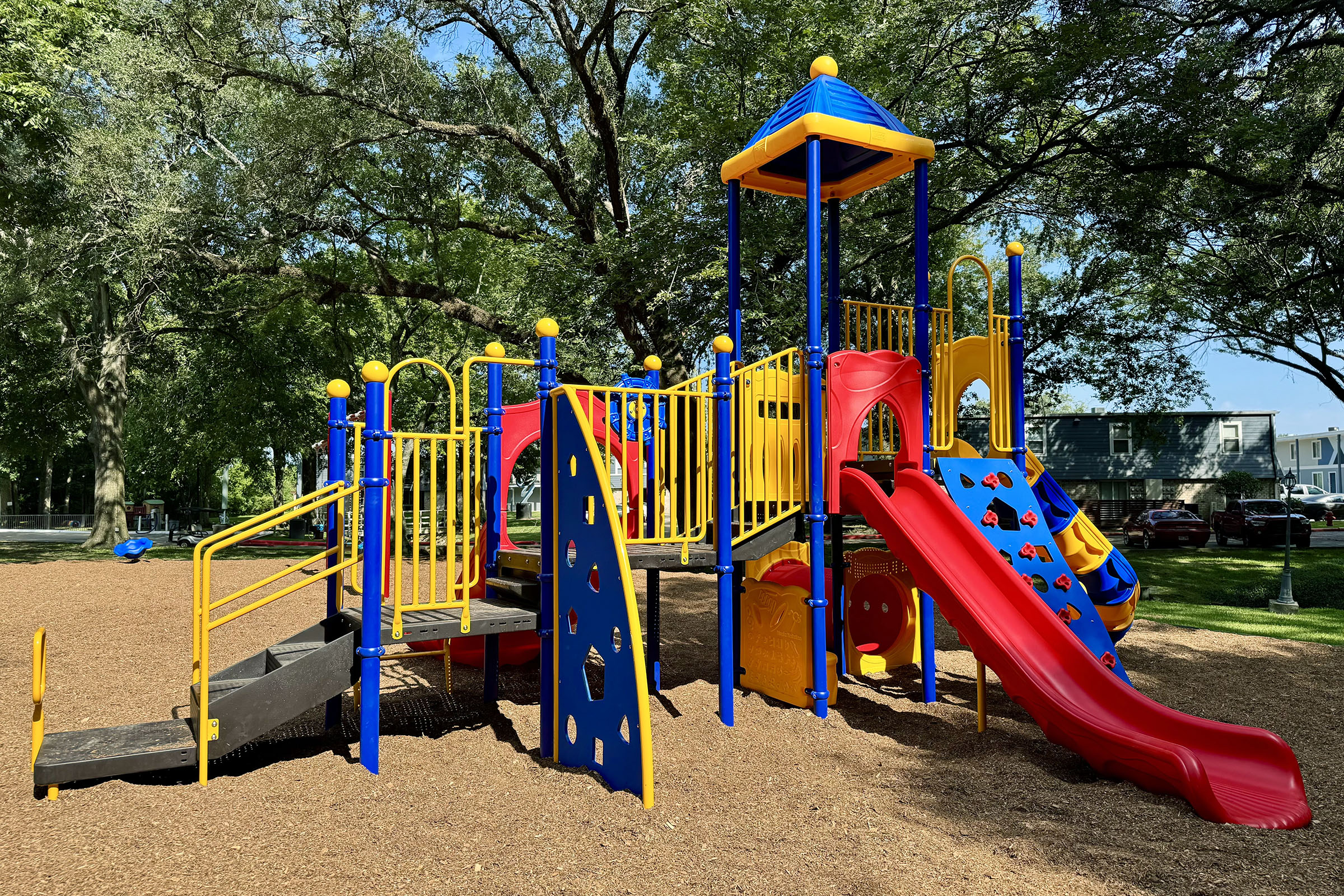A colorful playground with climbing structures, slides, and safety railings, surrounded by trees and a soft ground cover. The playground features yellow and blue equipment, including a tall tower, slides, and climbing walls, designed for children's play and recreation.