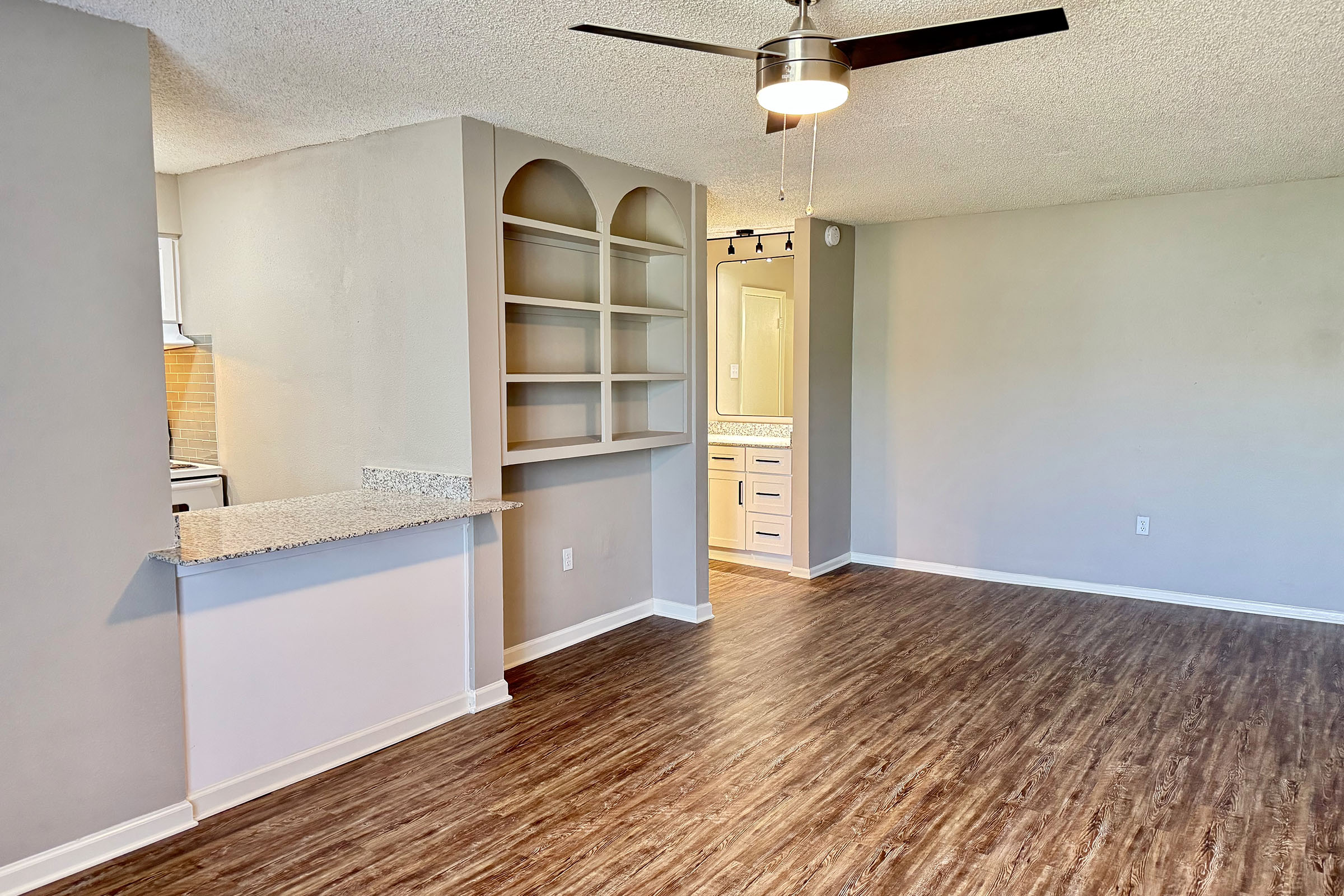 Bright and spacious living area with hardwood-style flooring. A wall-mounted fan hangs from the ceiling. On one side, a built-in bookshelf is visible, and on the other, there's an open kitchen area with light-colored cabinetry. The room has neutral walls, creating an inviting atmosphere.
