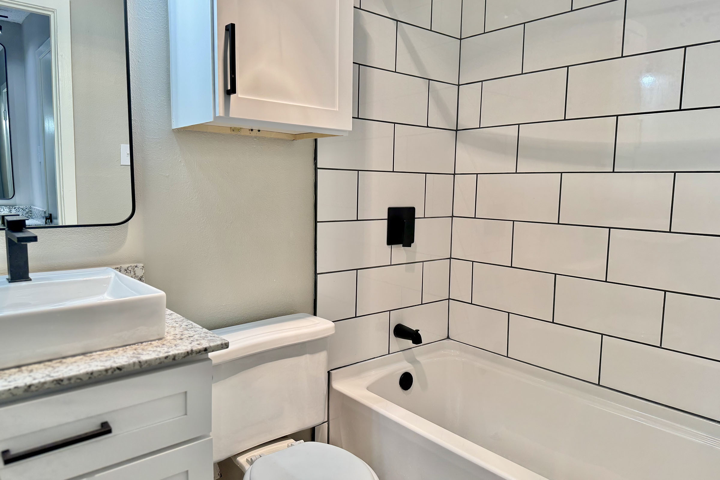 A modern bathroom featuring a white bathtub with a shower, black faucet, and white tile walls. There's a white vanity with a rectangular sink and a mirror above it. The room is well-lit and has a clean aesthetic with gray tones and crisp lines.