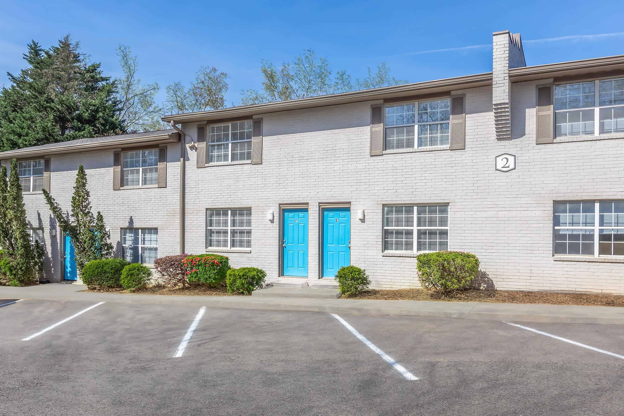 A two-story apartment building with a light brick exterior. The front features two doors painted blue, flanked by small, manicured shrubs. Windows with white frames are visible on both floors. The parking area in front has marked spaces and is bordered by greenery under a clear blue sky.