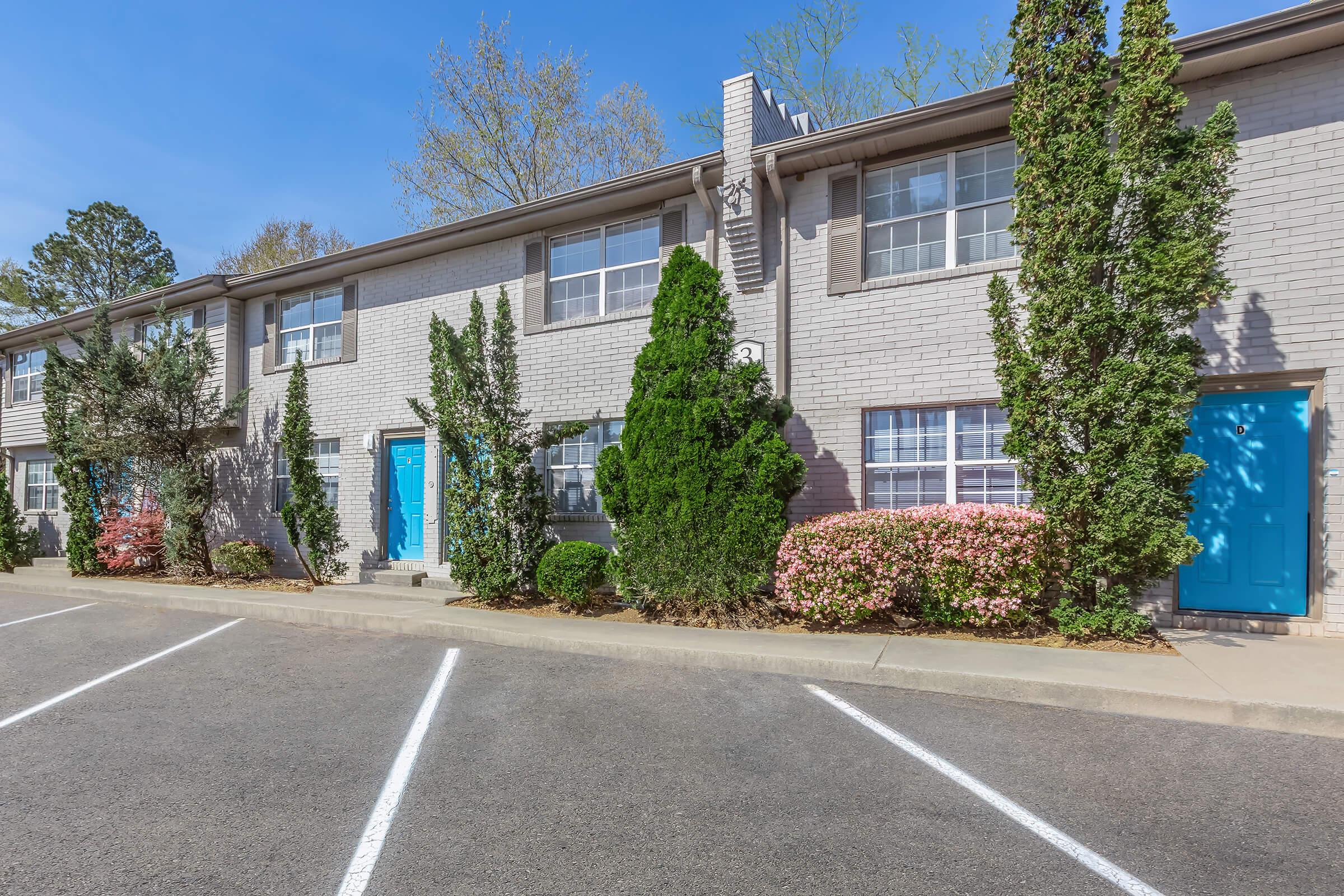 A row of residential apartment buildings with light gray brick exteriors, featuring blue front doors. Lush green shrubs and flowering plants are neatly arranged along the landscaped area. A paved parking lot with designated spaces is situated in front of the buildings, under a clear blue sky.