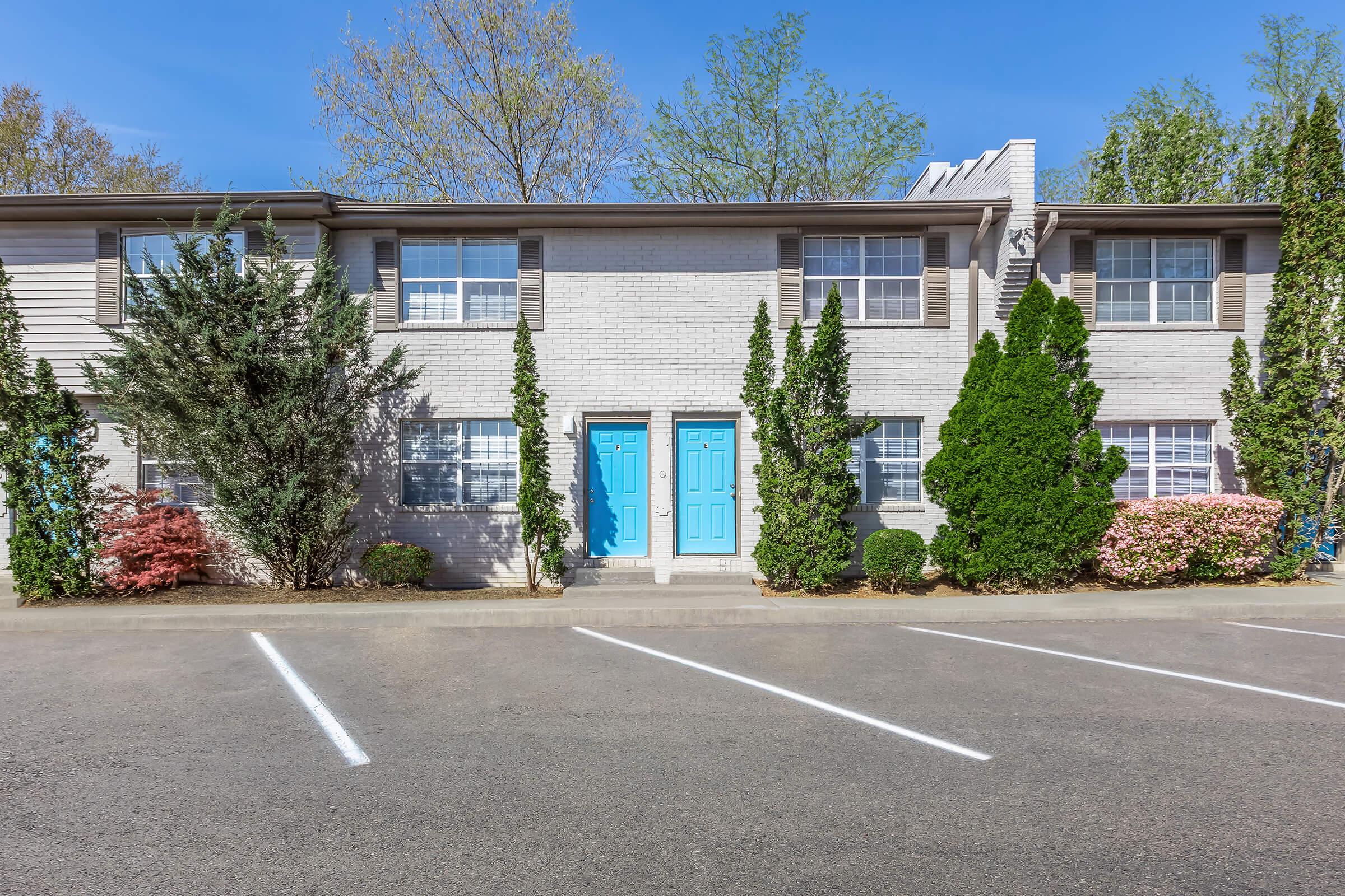 A two-story apartment building with light gray brick exterior and blue front doors, surrounded by neatly trimmed shrubs and flowering plants. The parking area in front features white striped lines and a clear blue sky overhead, with green trees in the background.