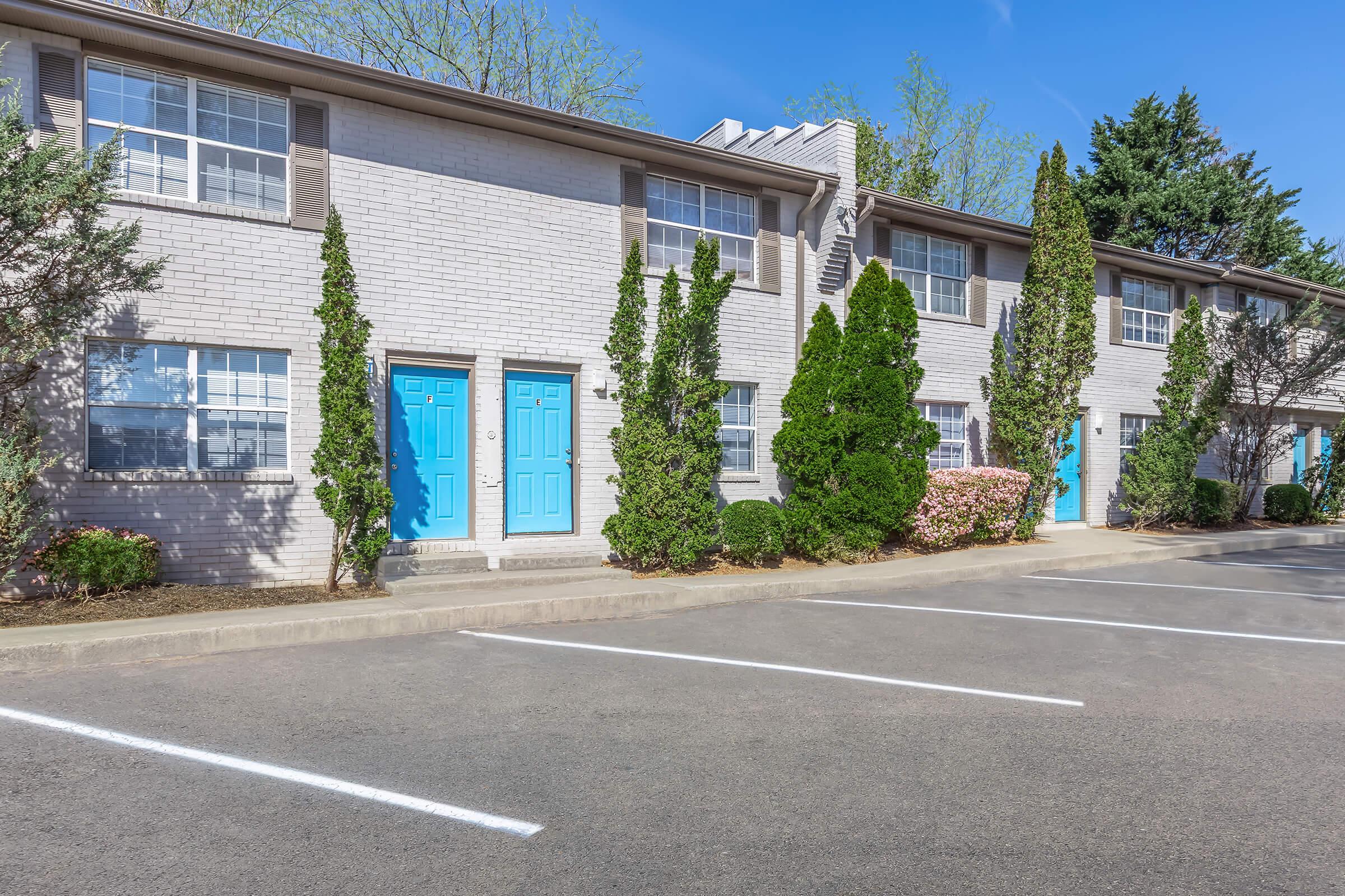 A row of two-story apartment buildings featuring light-colored brick exteriors and vibrant blue doors. The landscaping includes neatly trimmed shrubs and small trees, with a clear blue sky overhead. A parking area is visible in front, marked with white lines for vehicle spaces.