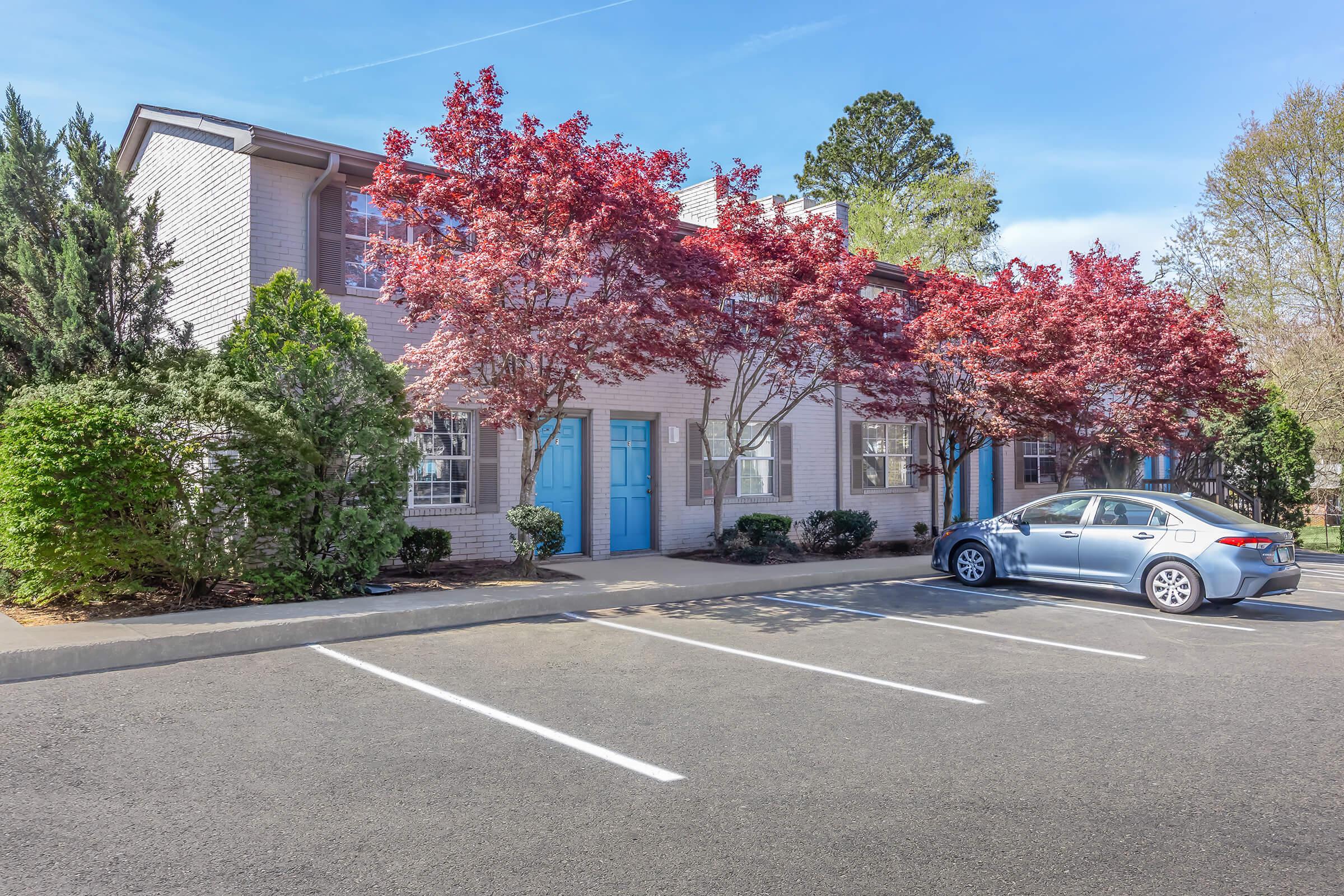 A residential building with light gray brick walls and blue doors. In front, there are vibrant red maple trees and a neatly paved parking lot with white lines. A silver car is parked beside the building, under a clear blue sky. Green shrubs line the walkway, adding to the inviting atmosphere.