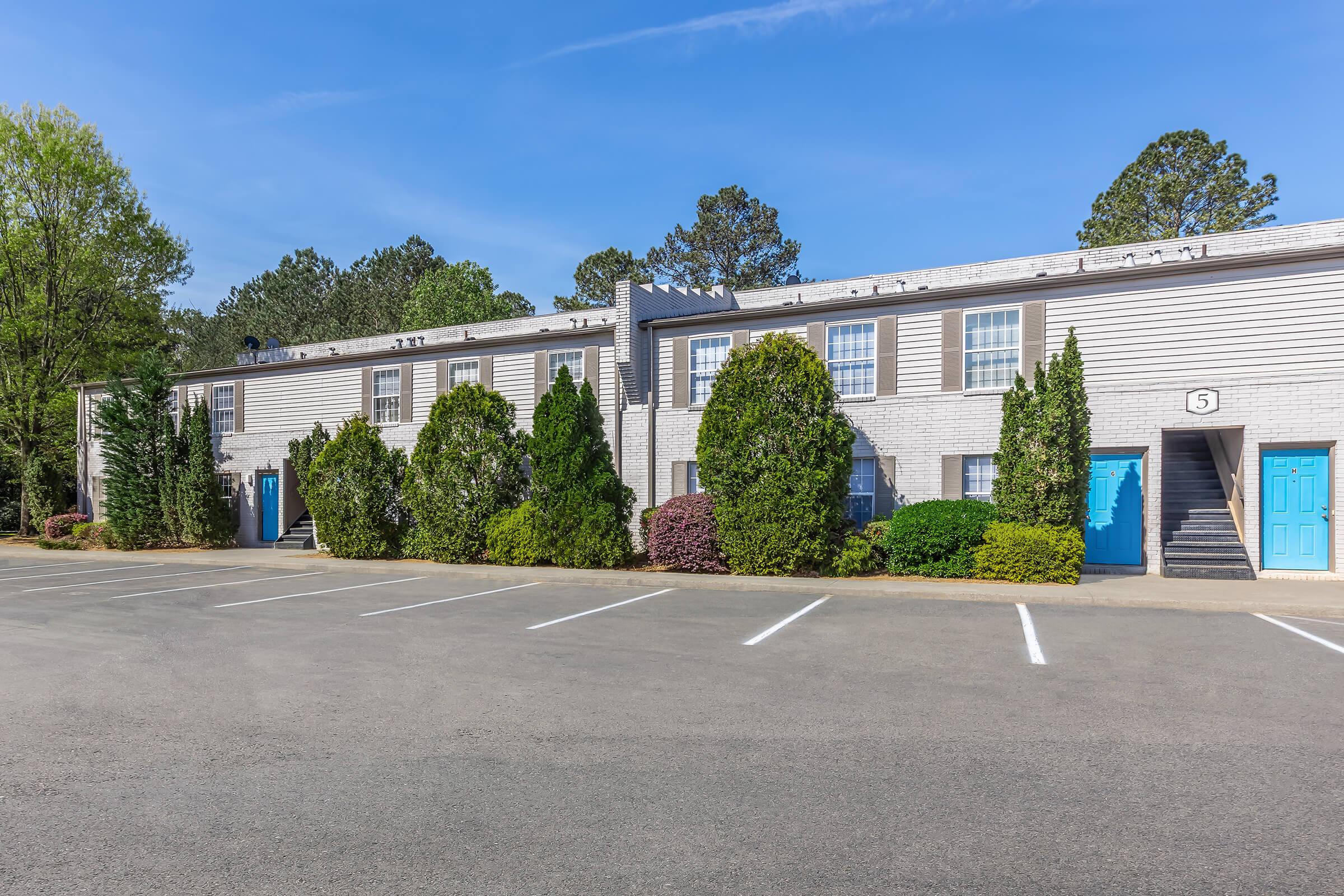 A view of a two-story apartment building with white siding and blue doors. Lush green shrubs and trees surround the entrance. The foreground features a spacious parking lot with clearly marked white parking spaces under a clear blue sky.