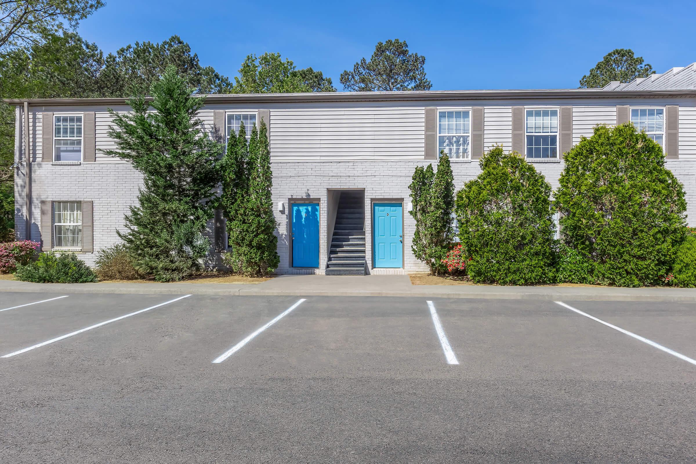 A two-story apartment building with light-colored siding and blue doors. The building is flanked by tall shrubs and trees, with a clear sky in the background. A parking area with marked spaces is visible in front of the building.