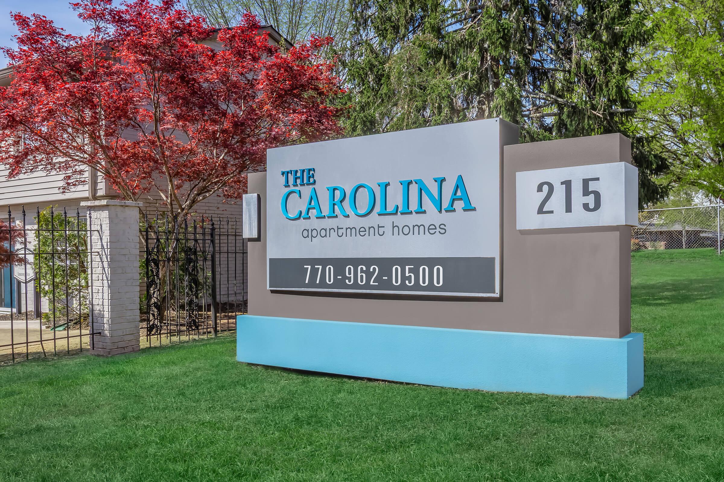 Sign for "The Carolina Apartment Homes" with the address "215" and phone number "770-962-0500." The sign is set on a grassy area with trees and a decorative red shrub in the background.