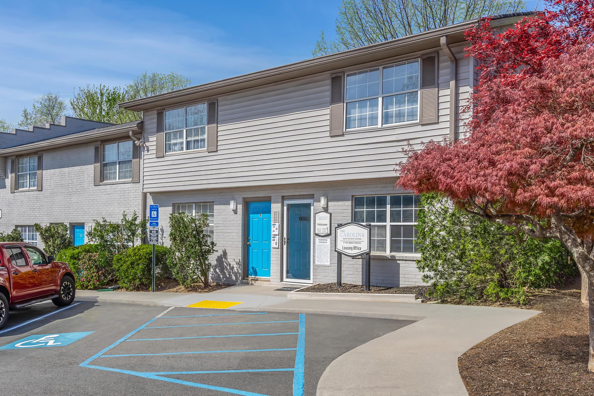 A modern, two-story residential building with light gray siding and blue front door. A red car is parked in the lot, and a vibrant red maple tree is in the foreground. The entrance features a sign with the building's name, surrounded by well-maintained greenery. Blue sky and trees are visible overhead.