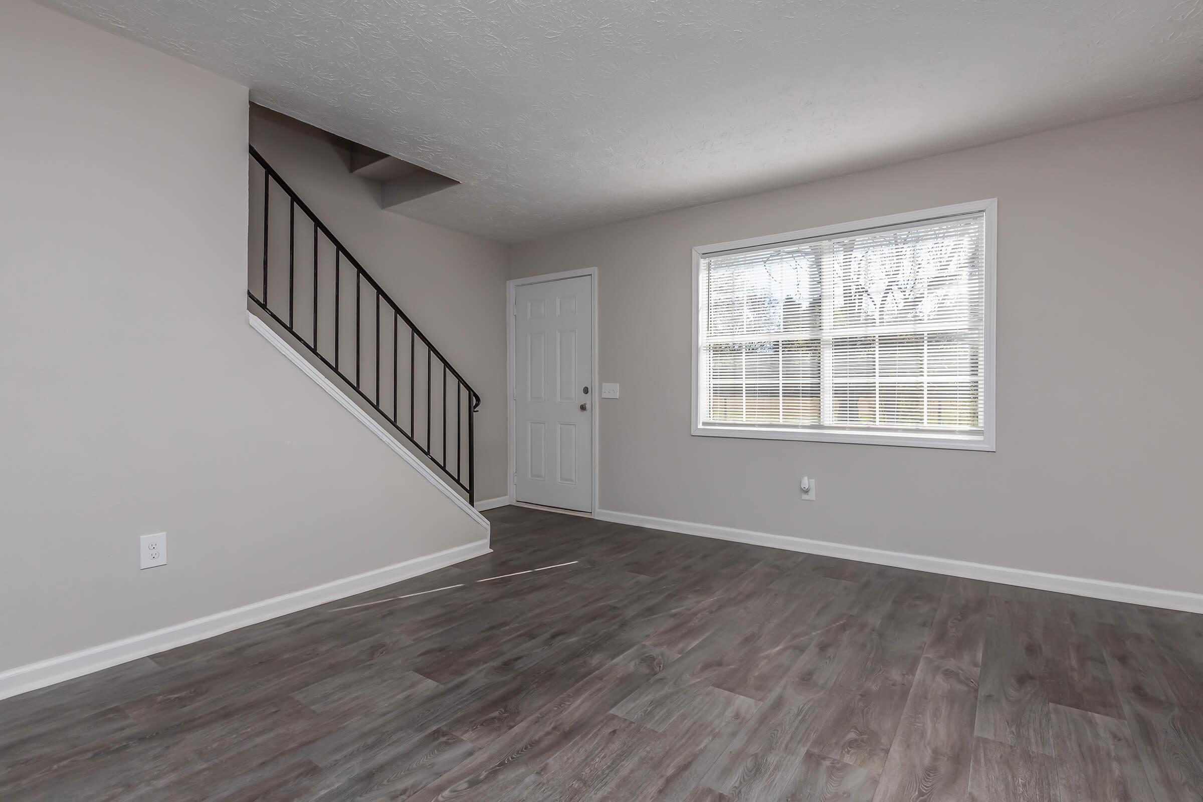 A vacant interior room with light gray walls and flooring. A staircase with a black railing is visible on the left side. A window with vertical blinds allows natural light to enter, highlighting the space. The room appears clean and well-maintained, suitable for various furniture arrangements.