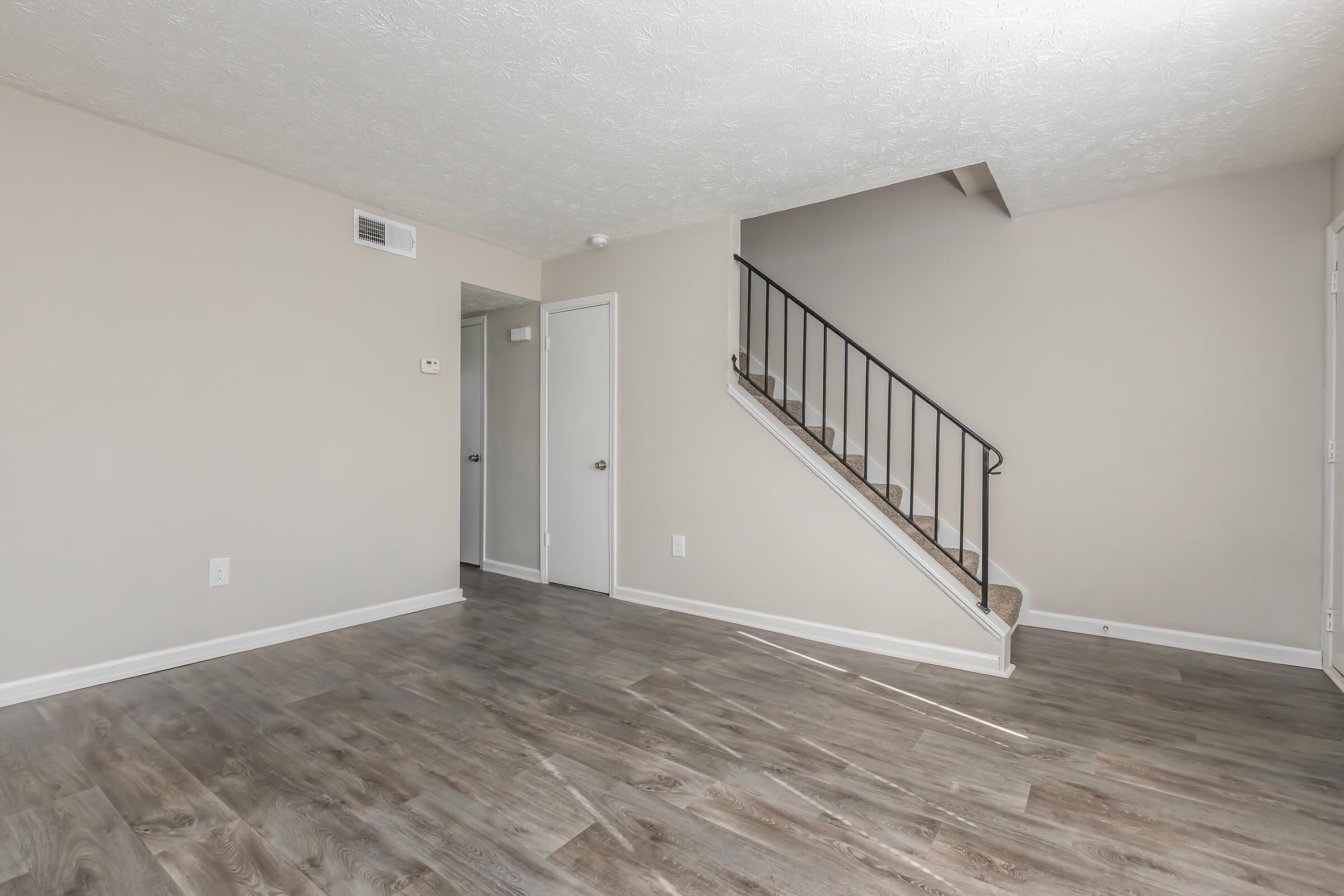 A spacious, minimally decorated room with light-colored walls and wood laminate flooring. A staircase with a black railing is visible in the background, and a doorway leads to another room. Natural light highlights the open space, creating a welcoming atmosphere.